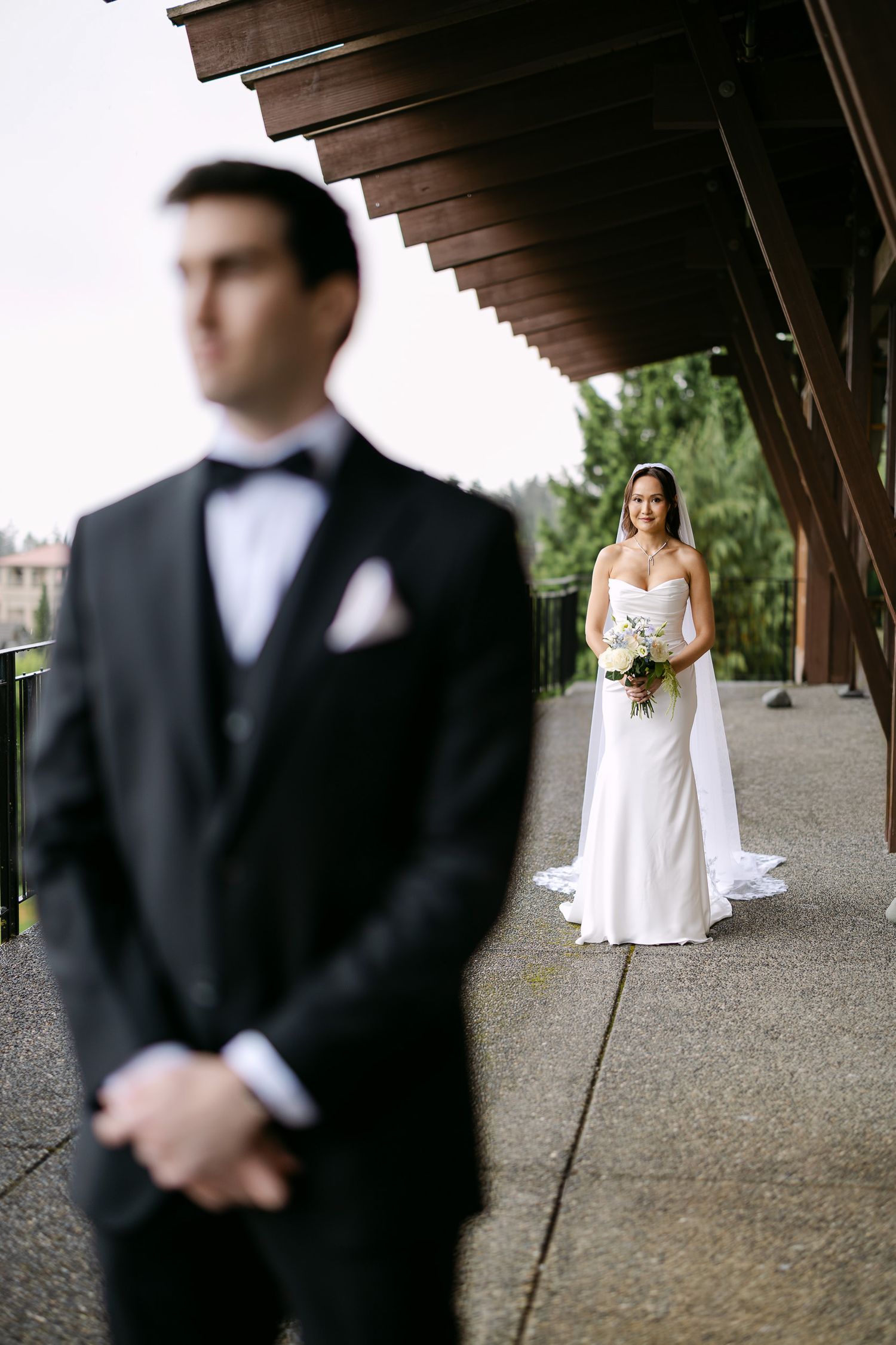 Bride in white gown holding bouquet stands behind groom in black tuxedo during elegant outdoor wedding portrait.