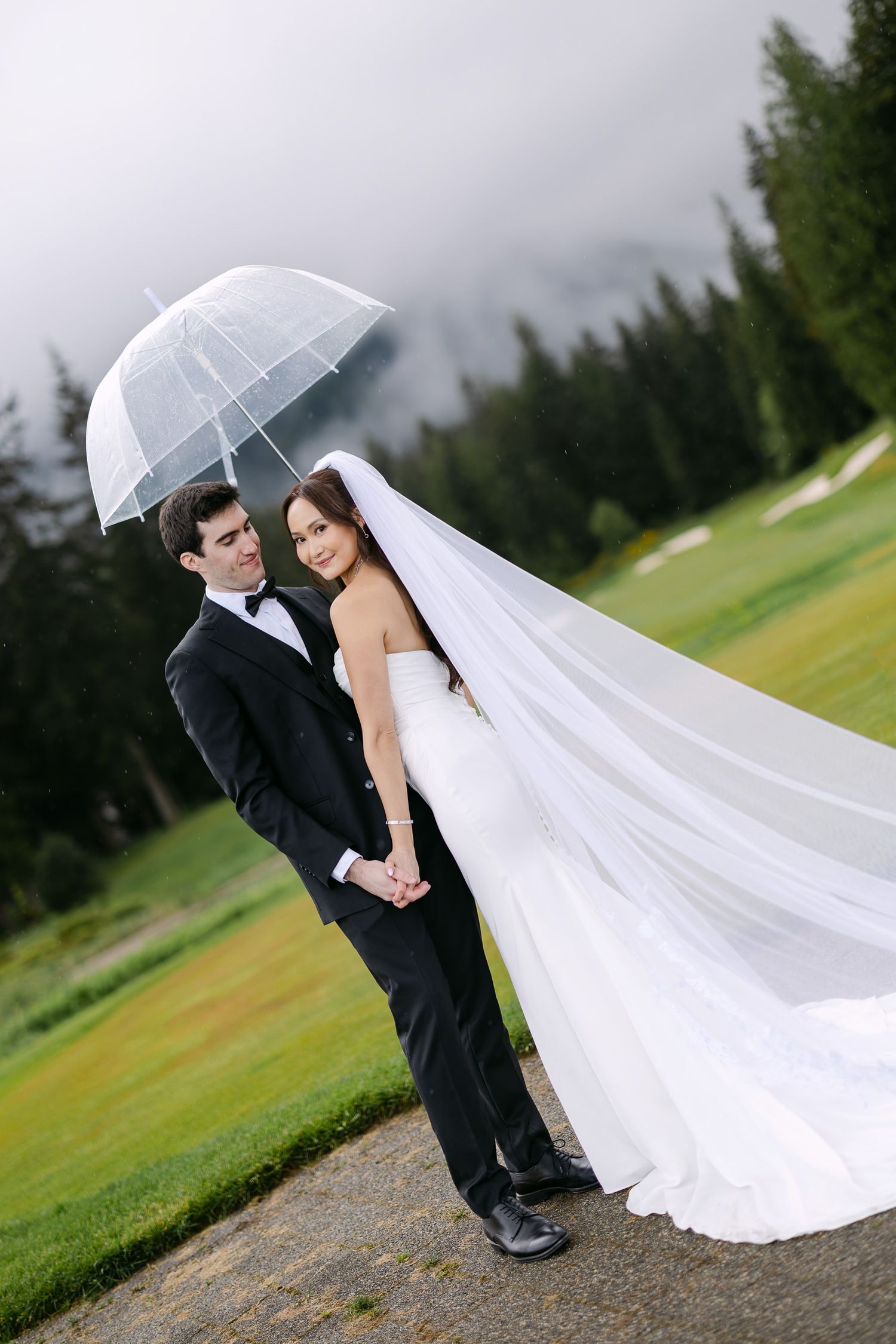 Bride and groom holding hands under a clear umbrella on a rainy wedding day with lush green trees in the background.