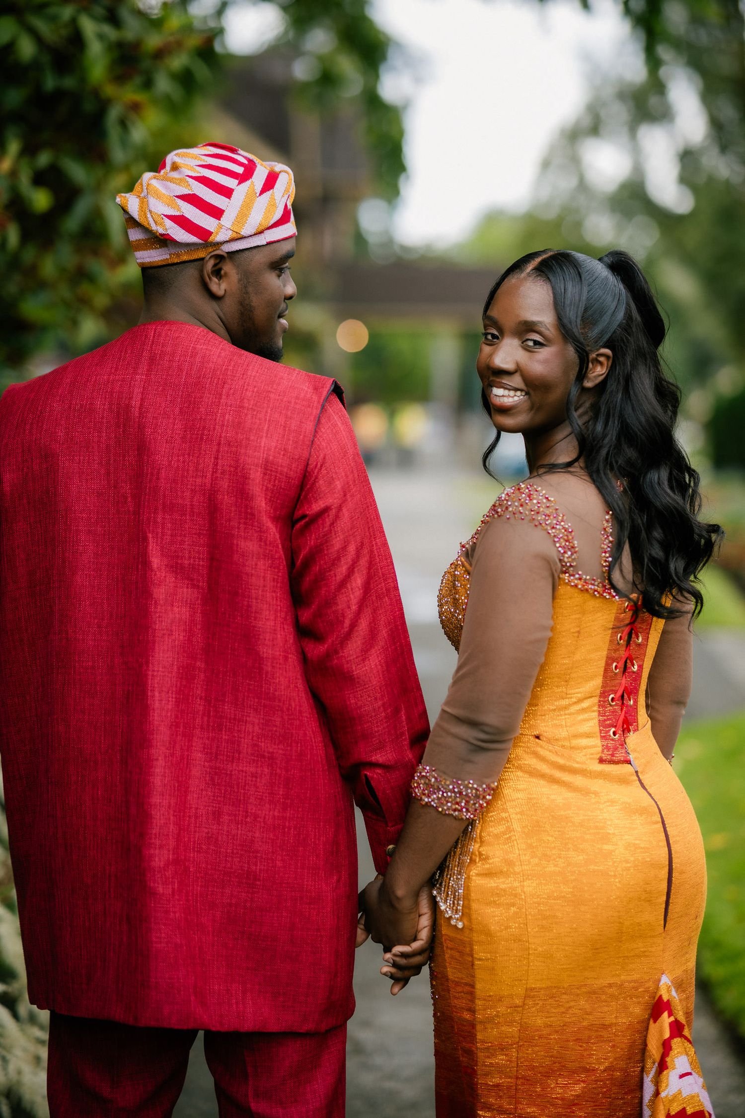 A couple in traditional African formal wear, with a red suit and orange dress, share a romantic moment outdoors.