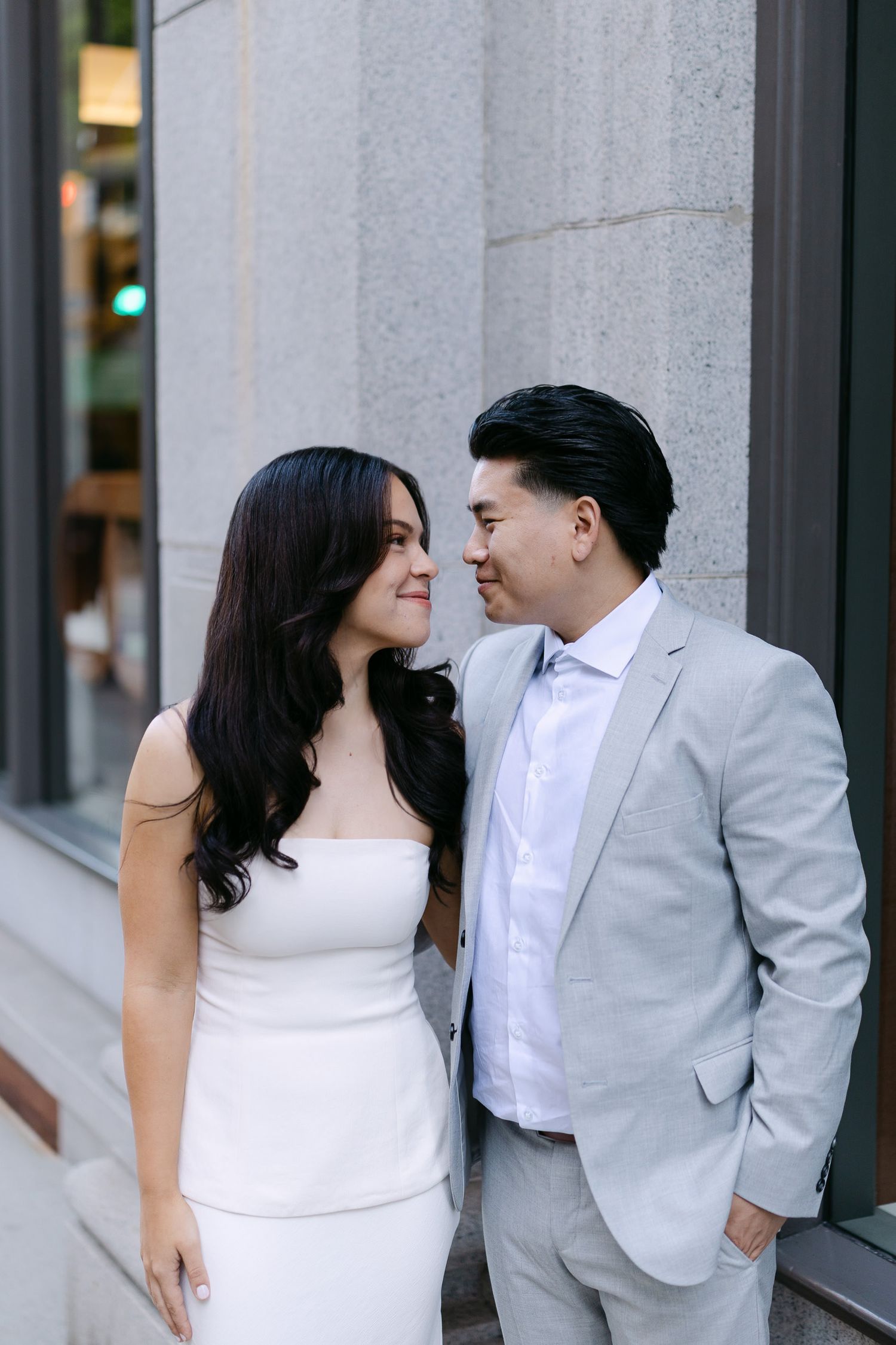 Smiling couple in a white dress and light gray suit stand nose to nose against a stone building for an engagement portrait.