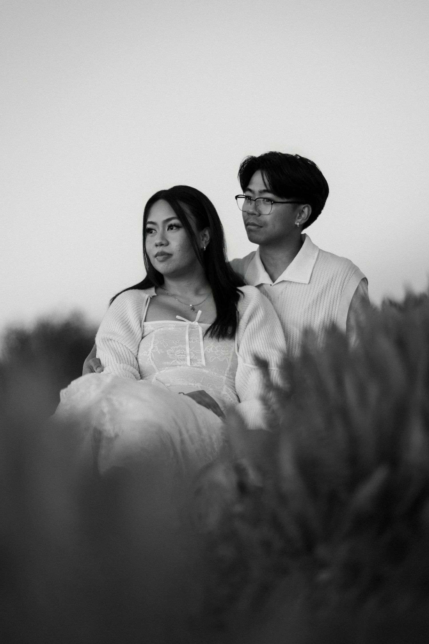 Black and white portrait of an engaged couple sitting close together, gazing into the distance behind soft blurred foliage.