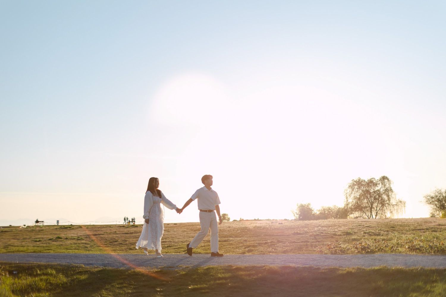 Engaged couple in white outfits walking hand in hand along a path through a field at a glowing sunset.