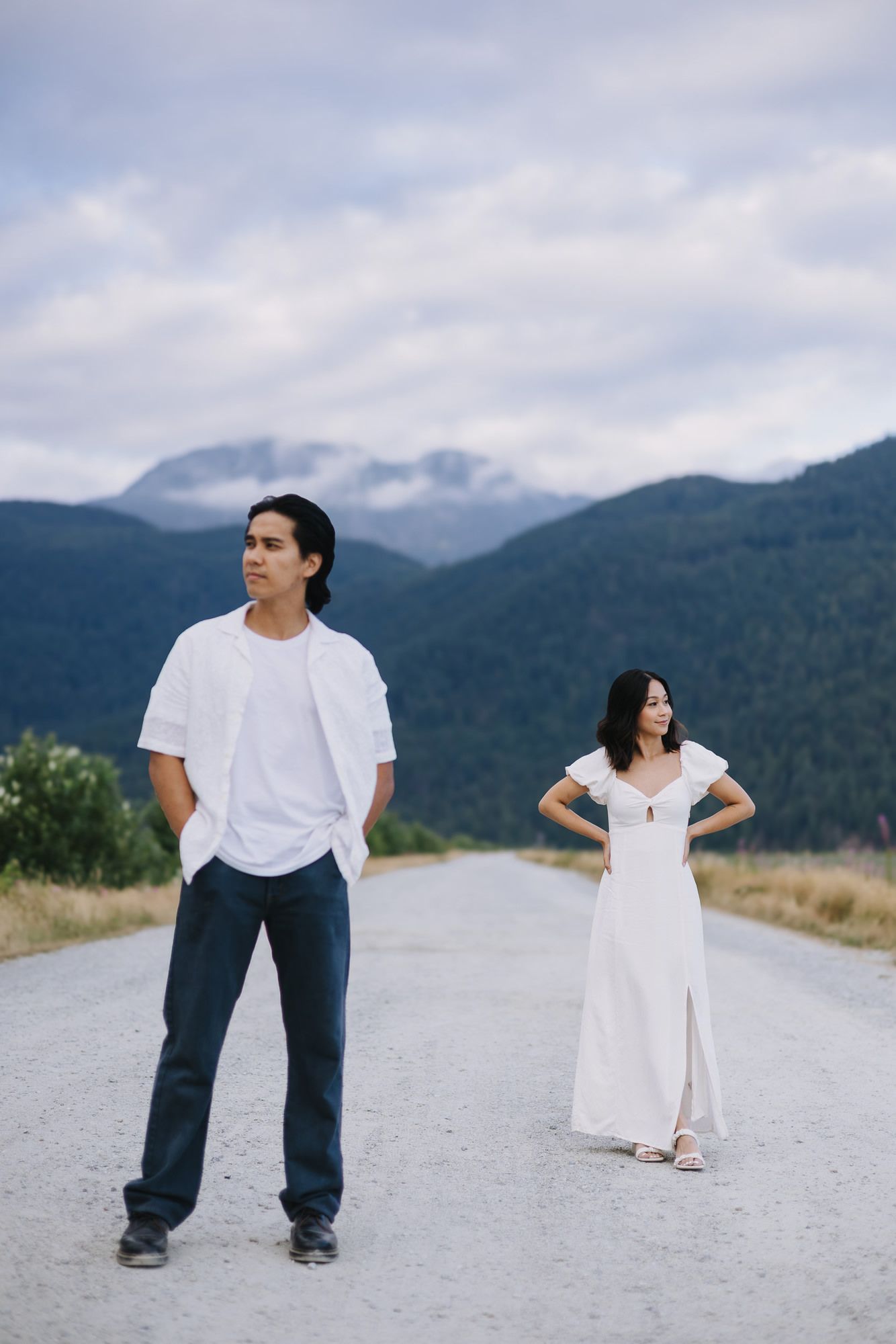 A man in a white shirt and a woman in a white dress stand apart on a gravel road with mountains behind them.