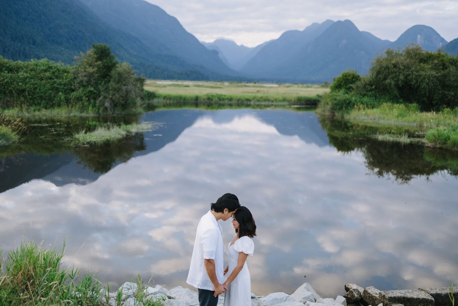Couple in white embracing at the edge of a calm lake that mirrors the cloudy sky, with green mountains in the background.