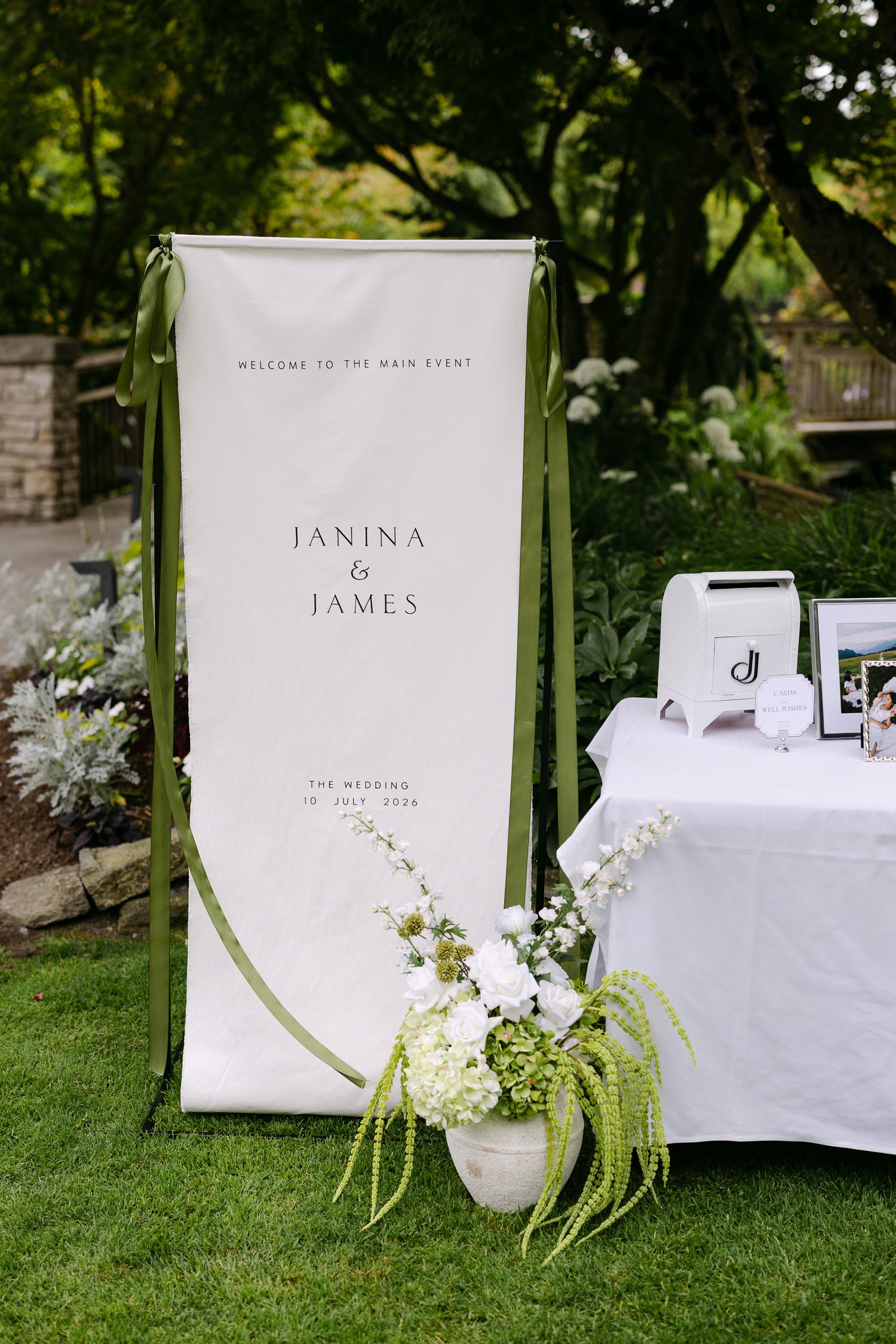 Outdoor wedding welcome banner for Janina and James with green ribbon accents and white floral arrangement on lush garden lawn.