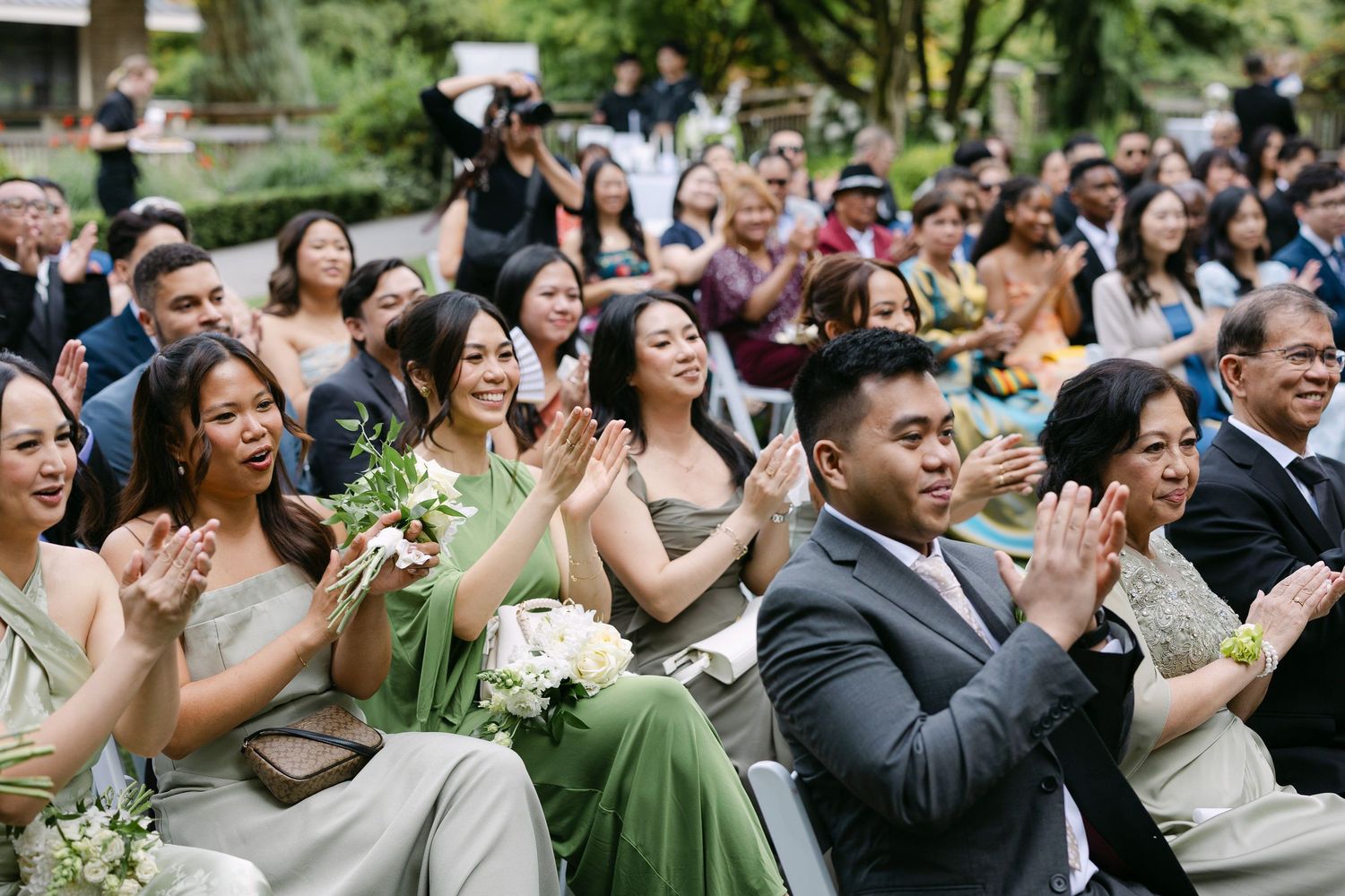 Wedding guests in sage green bridesmaid dresses and suits clapping joyfully during an outdoor garden ceremony.