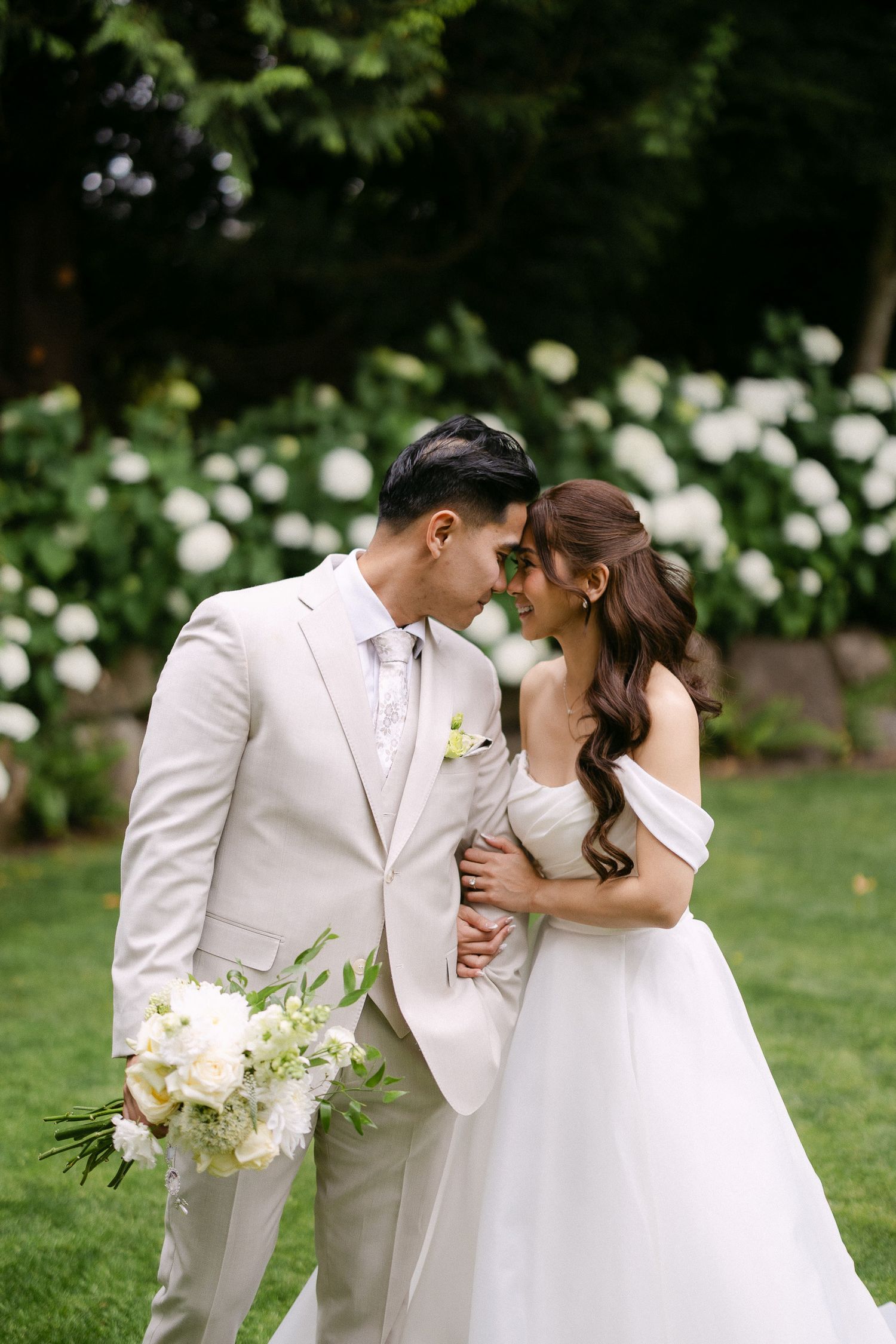 Bride and groom in white attire share a romantic moment outdoors, surrounded by lush greenery and white hydrangeas.