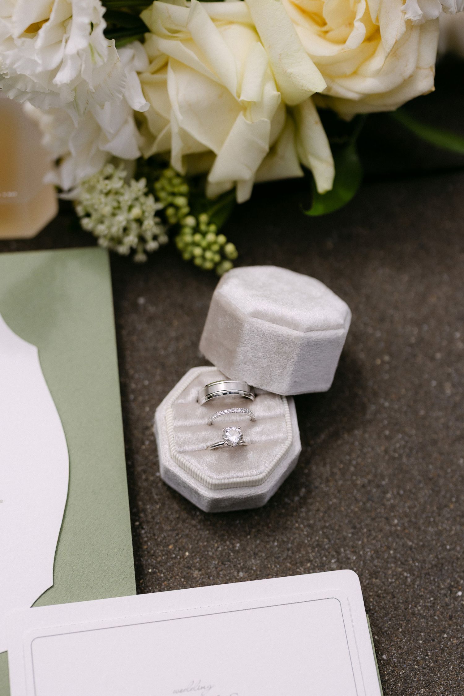 Wedding rings displayed in a gray velvet octagonal ring box beside white roses and elegant stationery.