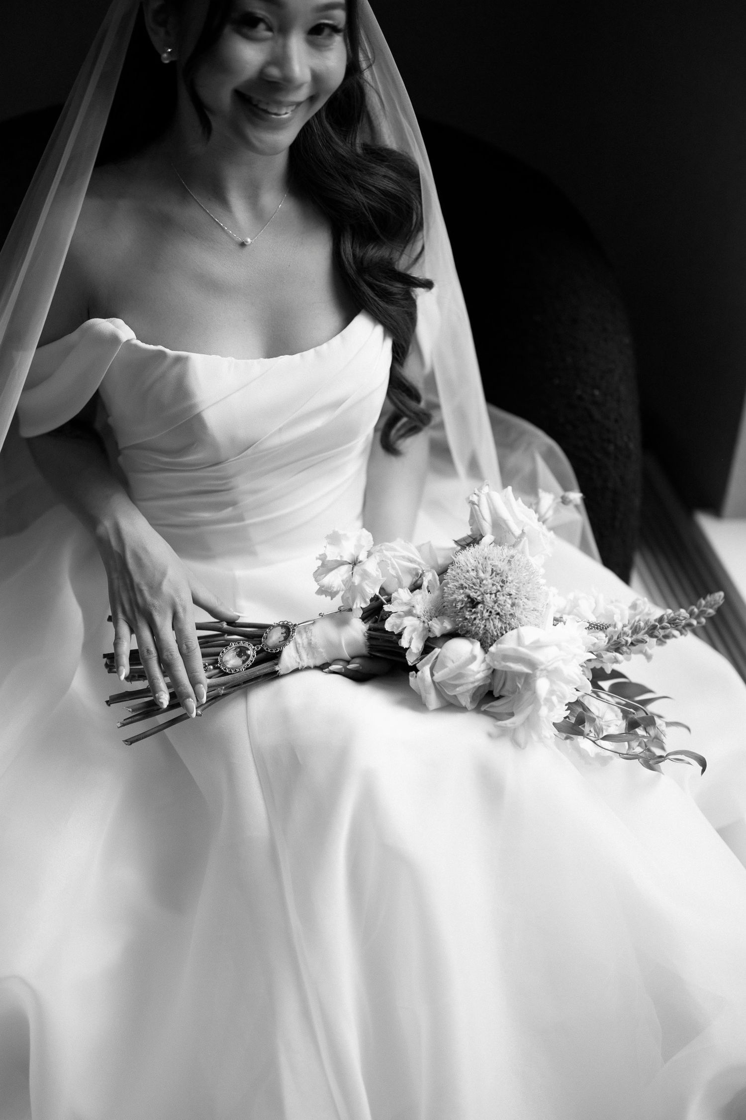 Black and white photo of a smiling bride in a strapless gown holding a floral bouquet while seated.