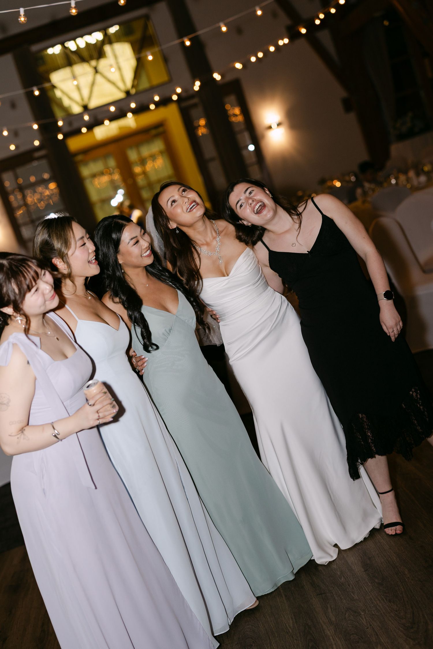 Bride in white gown laughing with bridesmaids in pastel dresses on wedding reception dance floor.