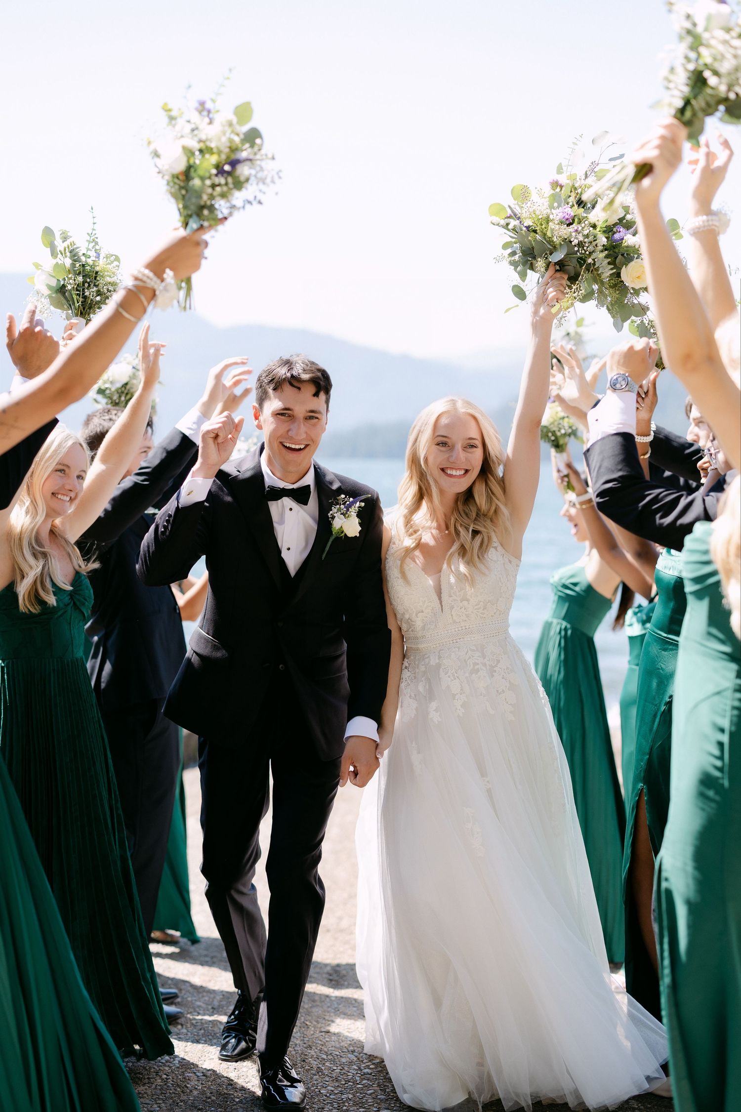 Bride and groom walk through cheering wedding party in green dresses by a scenic lake.