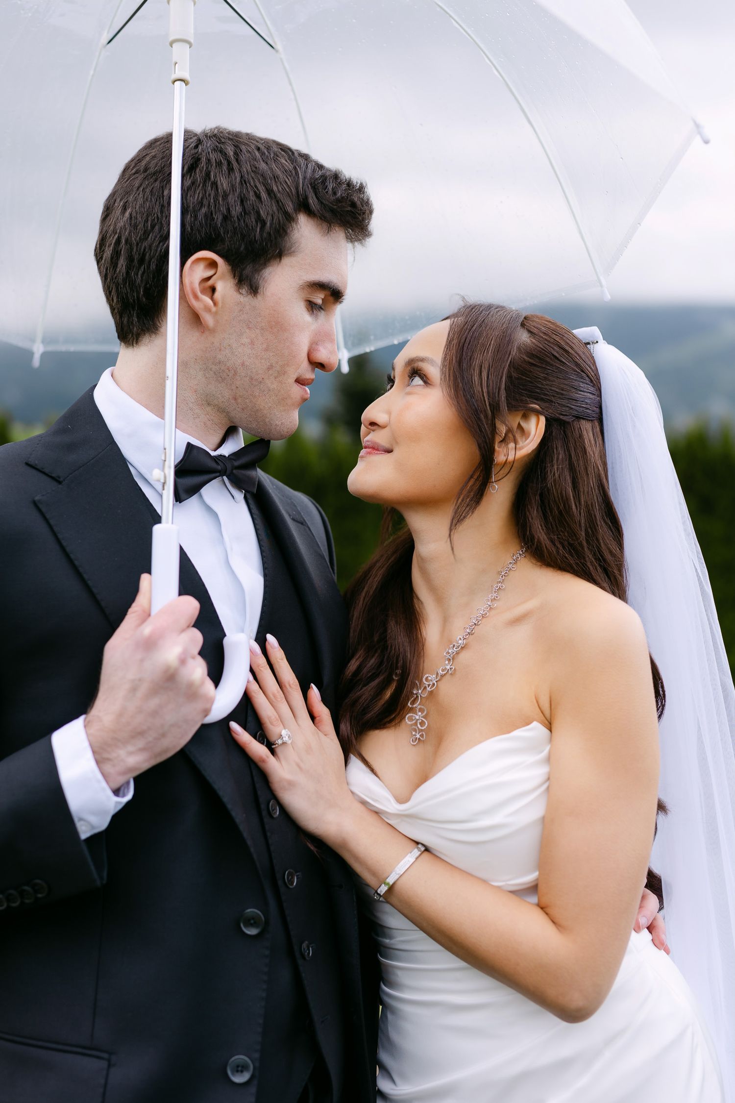 Bride and groom share a romantic moment under a clear umbrella on their rainy wedding day.