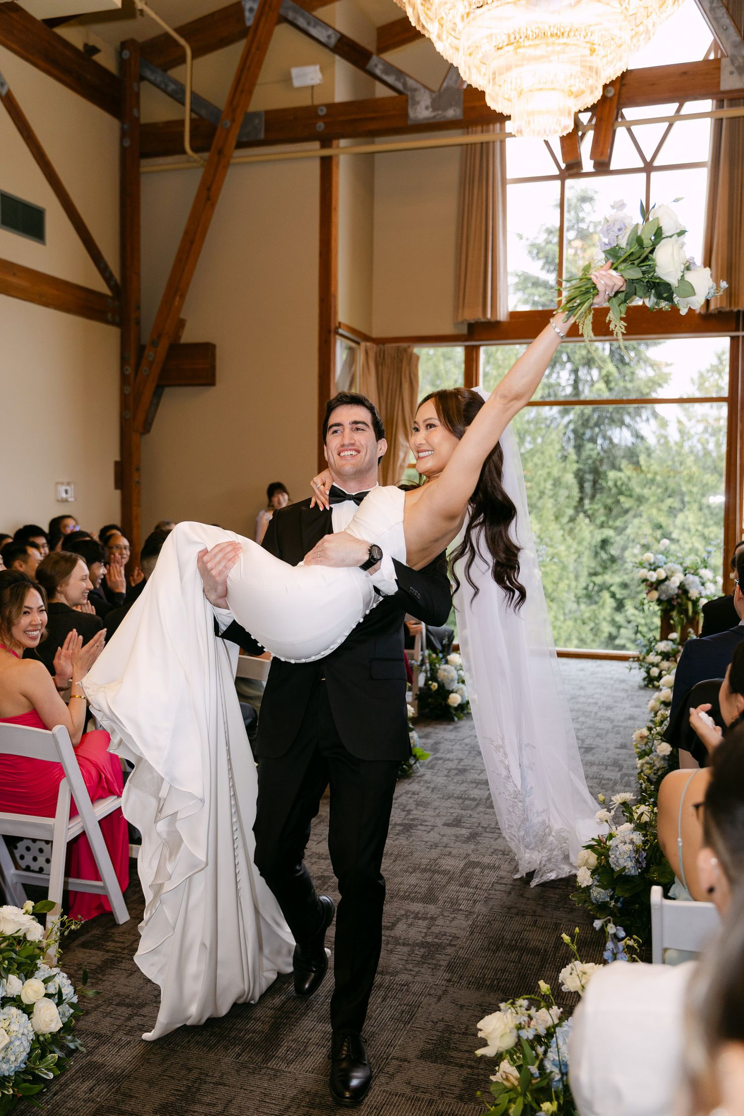 Groom carries bride down the aisle in elegant wedding venue, bride raises bouquet in celebration.