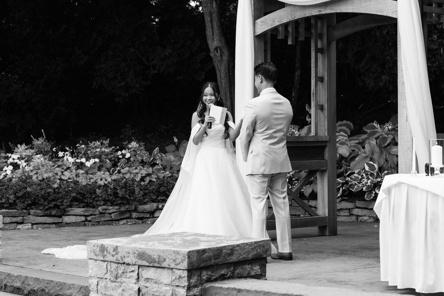Black and white photo of bride reading vows to groom at outdoor garden ceremony.
