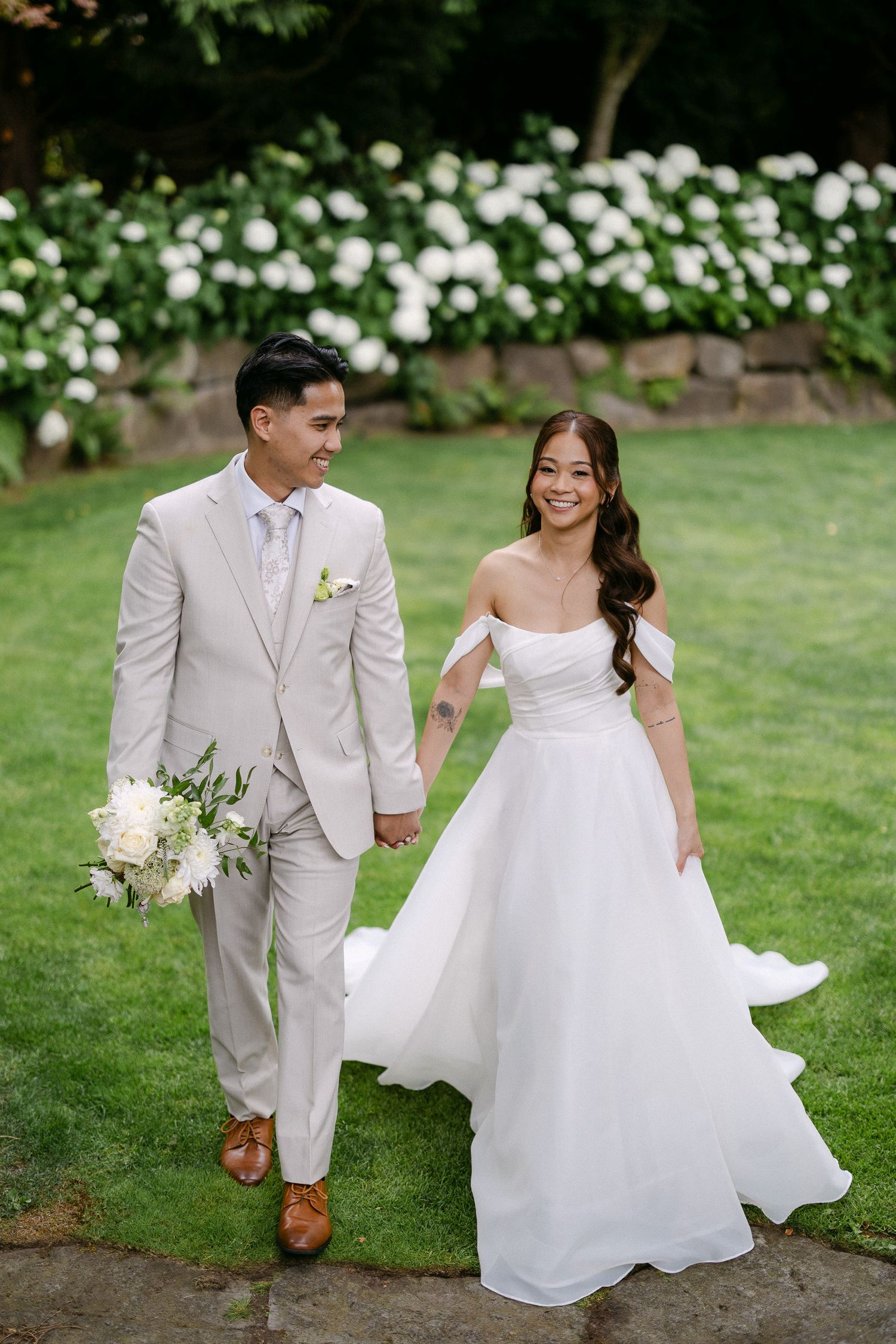 Bride in off-shoulder white gown and groom in beige suit hold hands walking on green lawn with white hydrangeas.