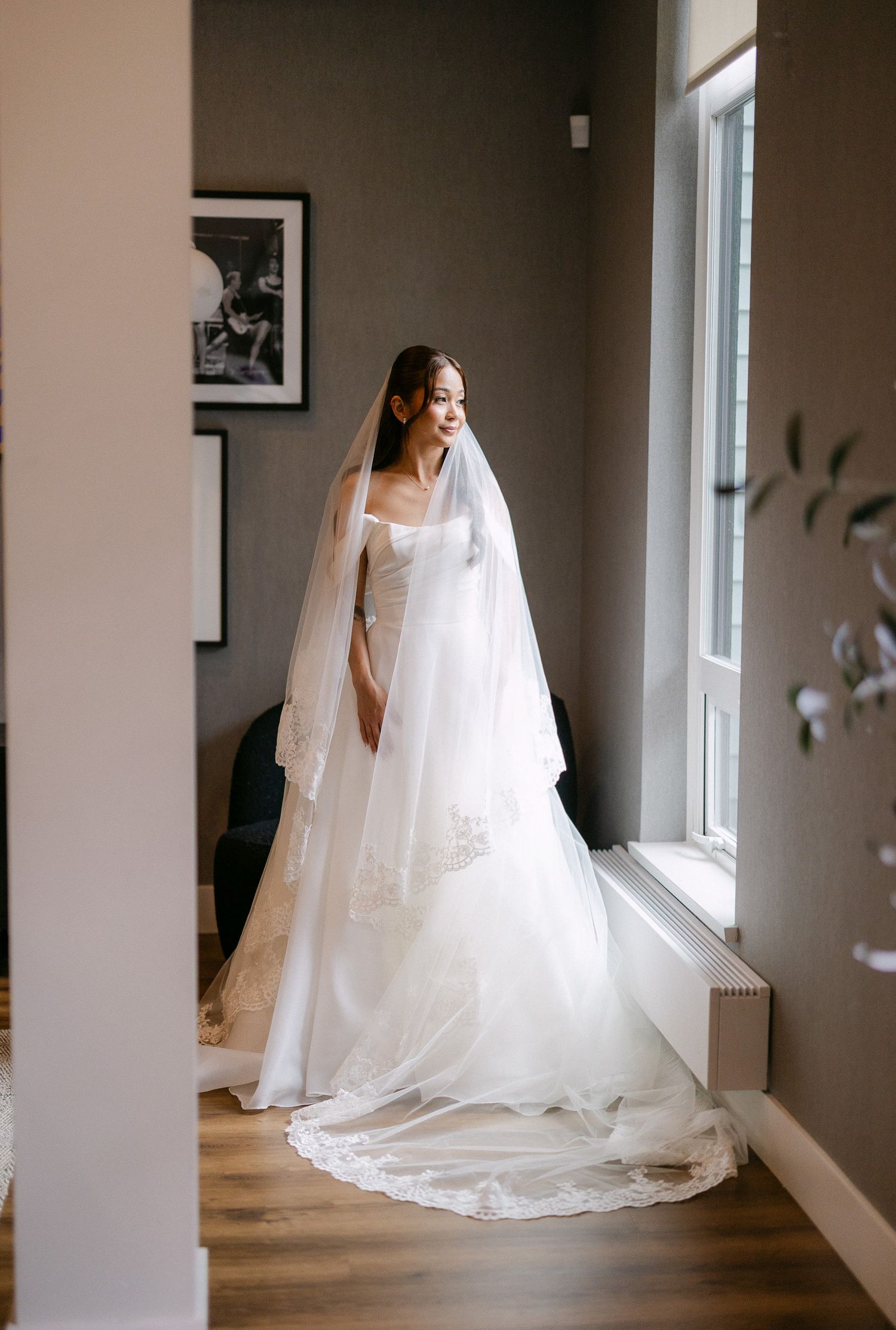 Bride in white strapless ball gown with lace cathedral veil standing by window in elegant bridal suite.