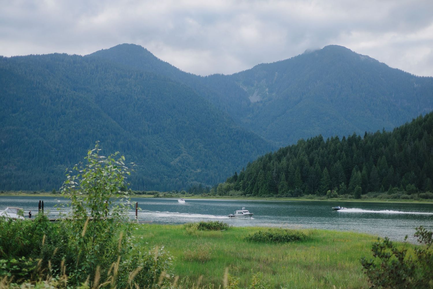 Calm mountain lake surrounded by forested peaks and green grassy shoreline under a cloudy sky, with small boats on the water.