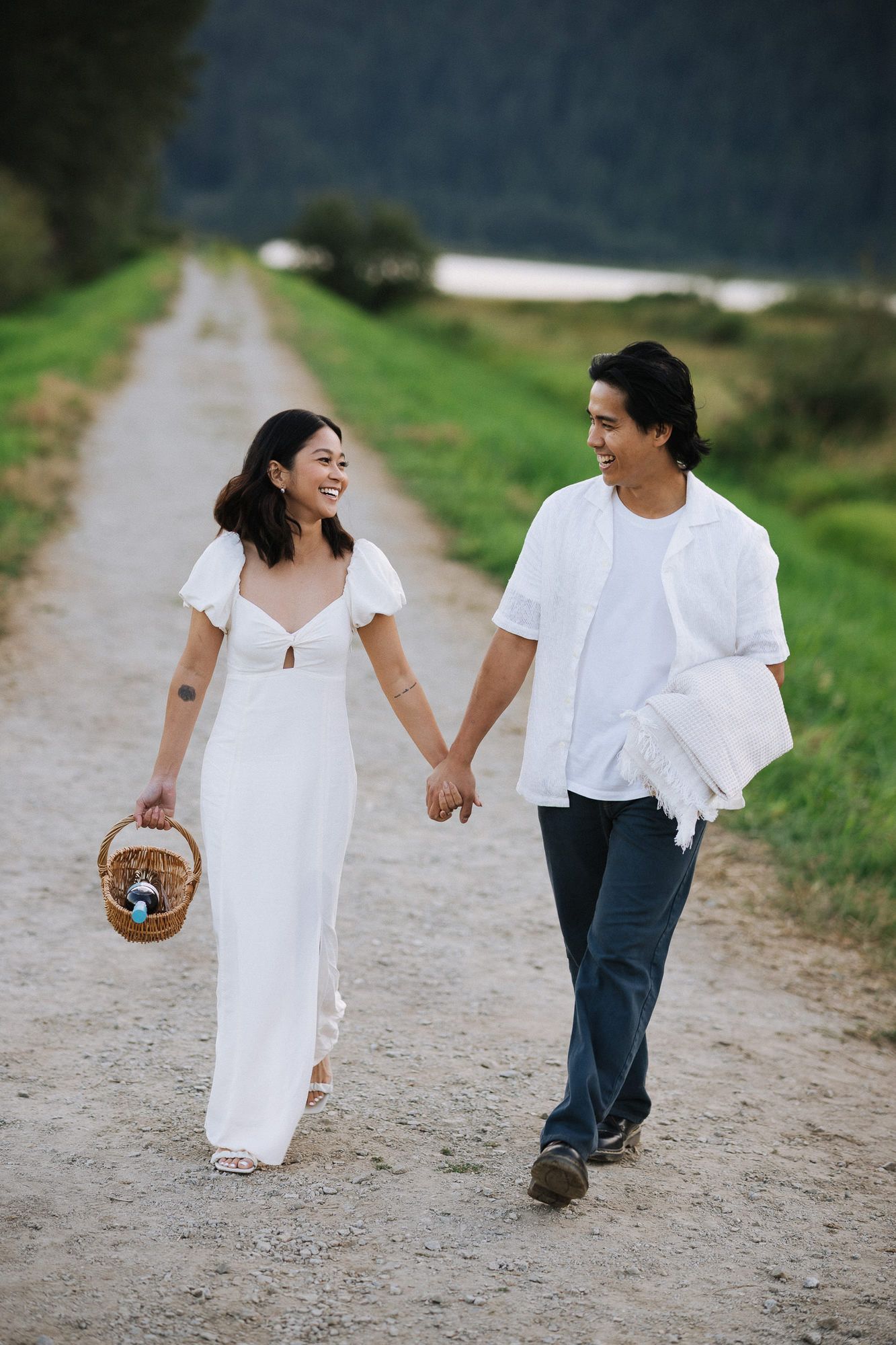 Smiling couple holding hands and walking along a gravel path with a picnic basket, surrounded by green grass and mountains.