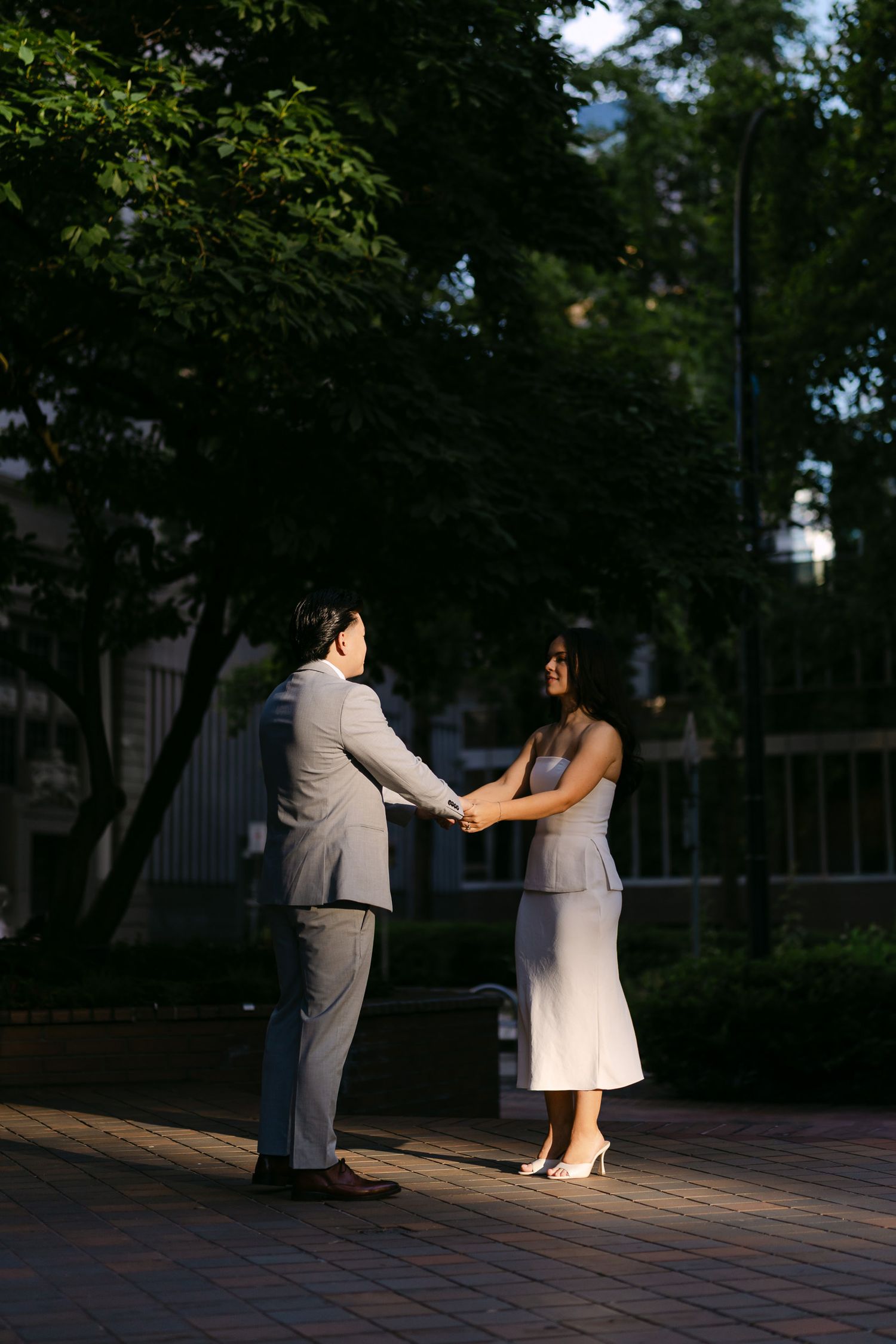 Couple in gray suit and white dress hold hands facing each other in a sunlit, tree-lined city plaza.