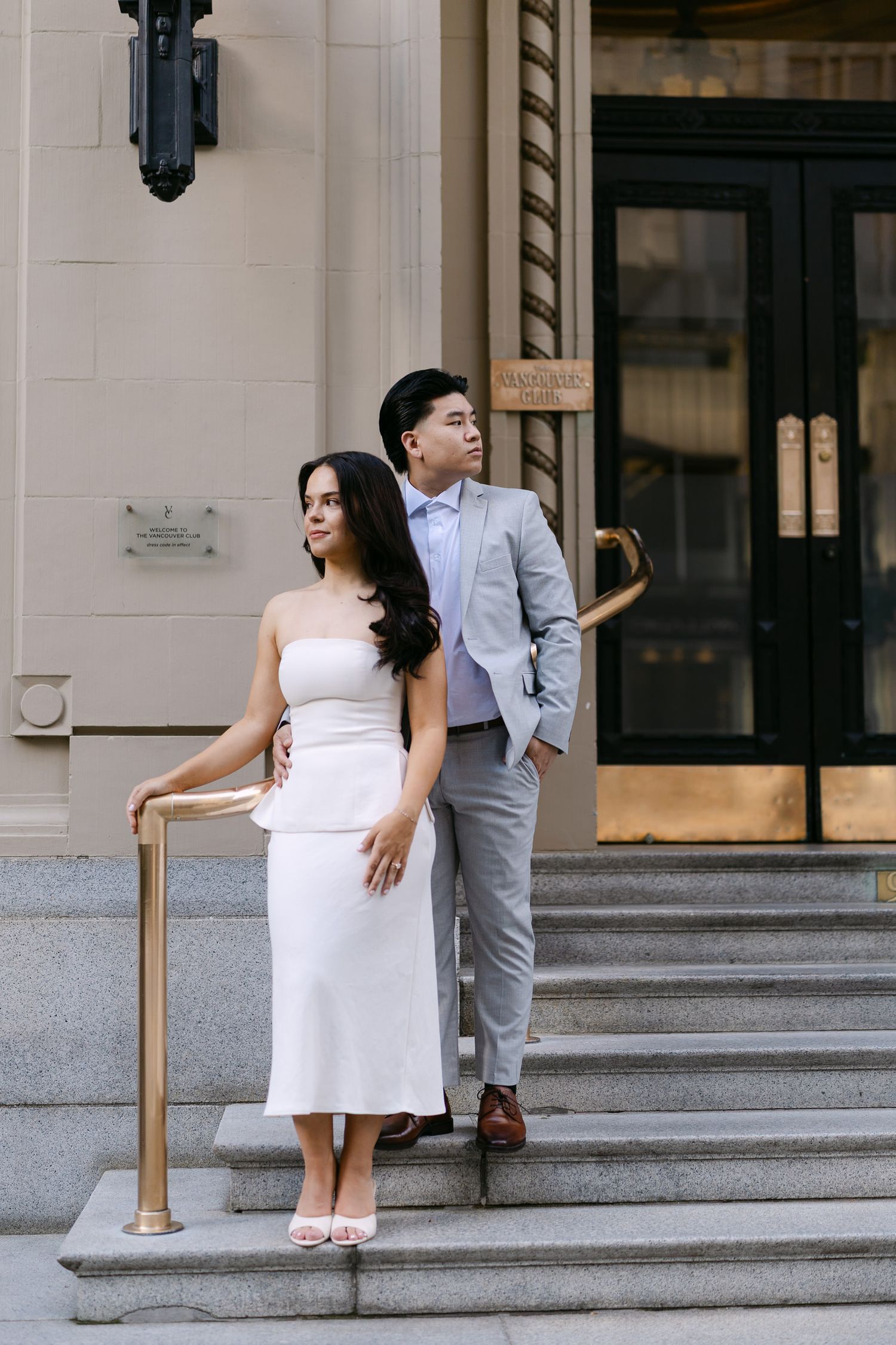 Couple stand on stone steps with brass handrails, the woman in white and the man in a gray suit, near a grand building entrance.