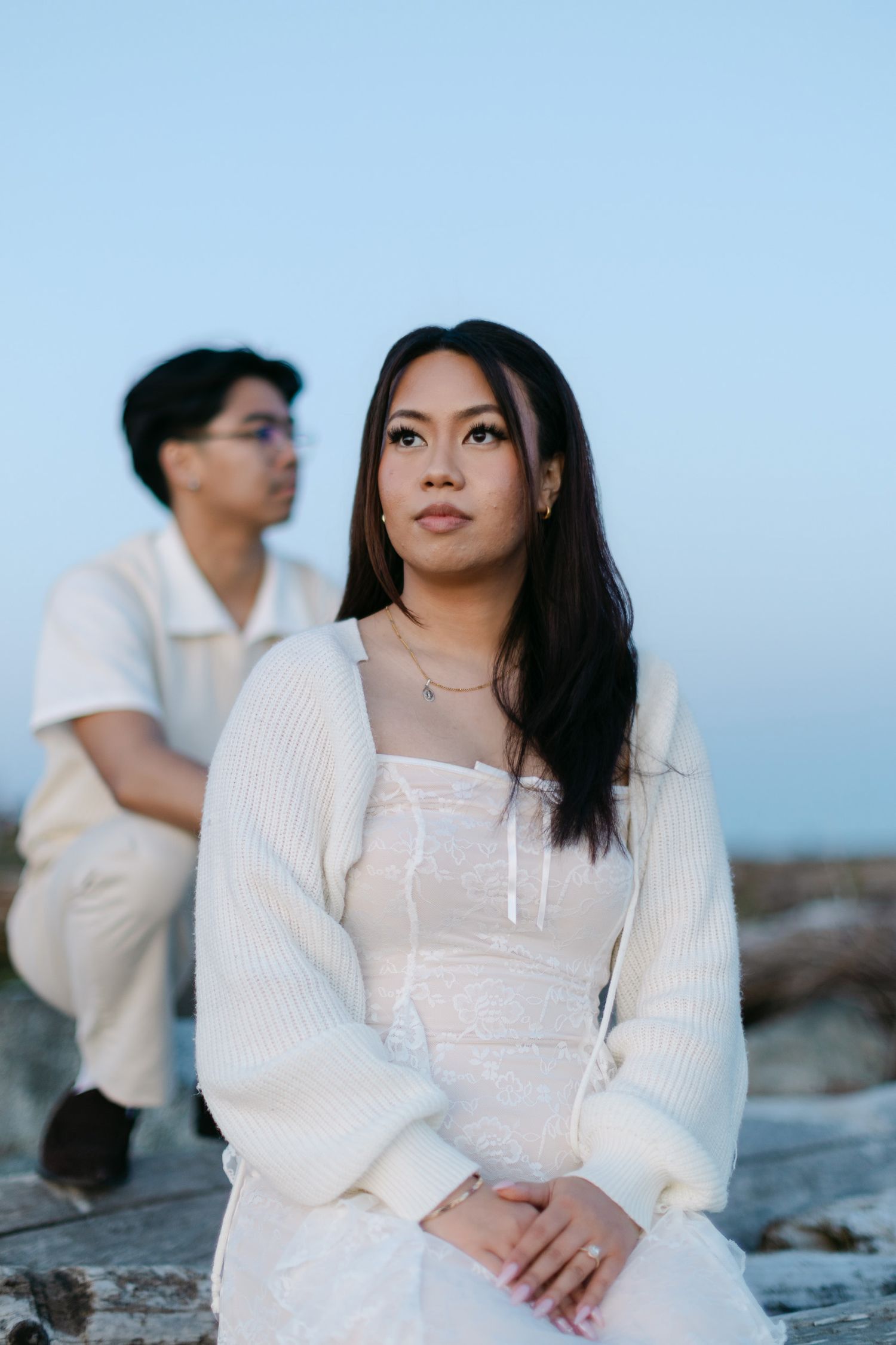 Woman in a white knit cardigan and dress gazes into the distance while a man in glasses sits blurred behind her against a soft blue sky.