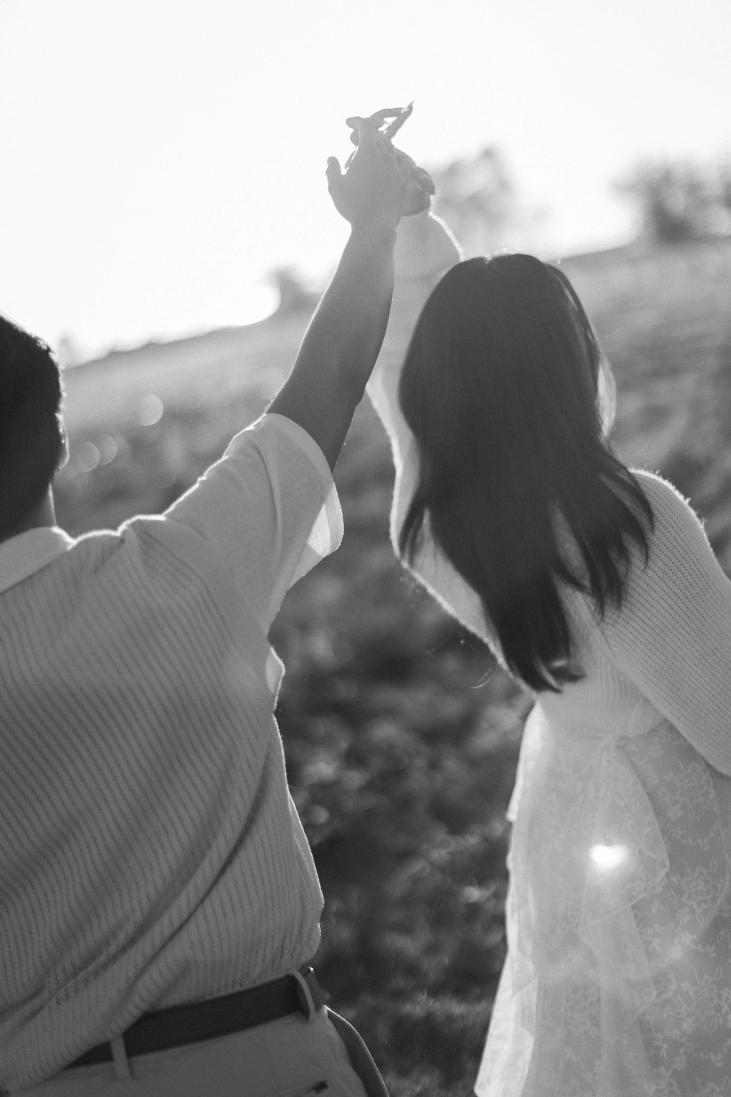 Black and white photo of a couple from behind holding hands raised high while walking through a sunlit grassy field.