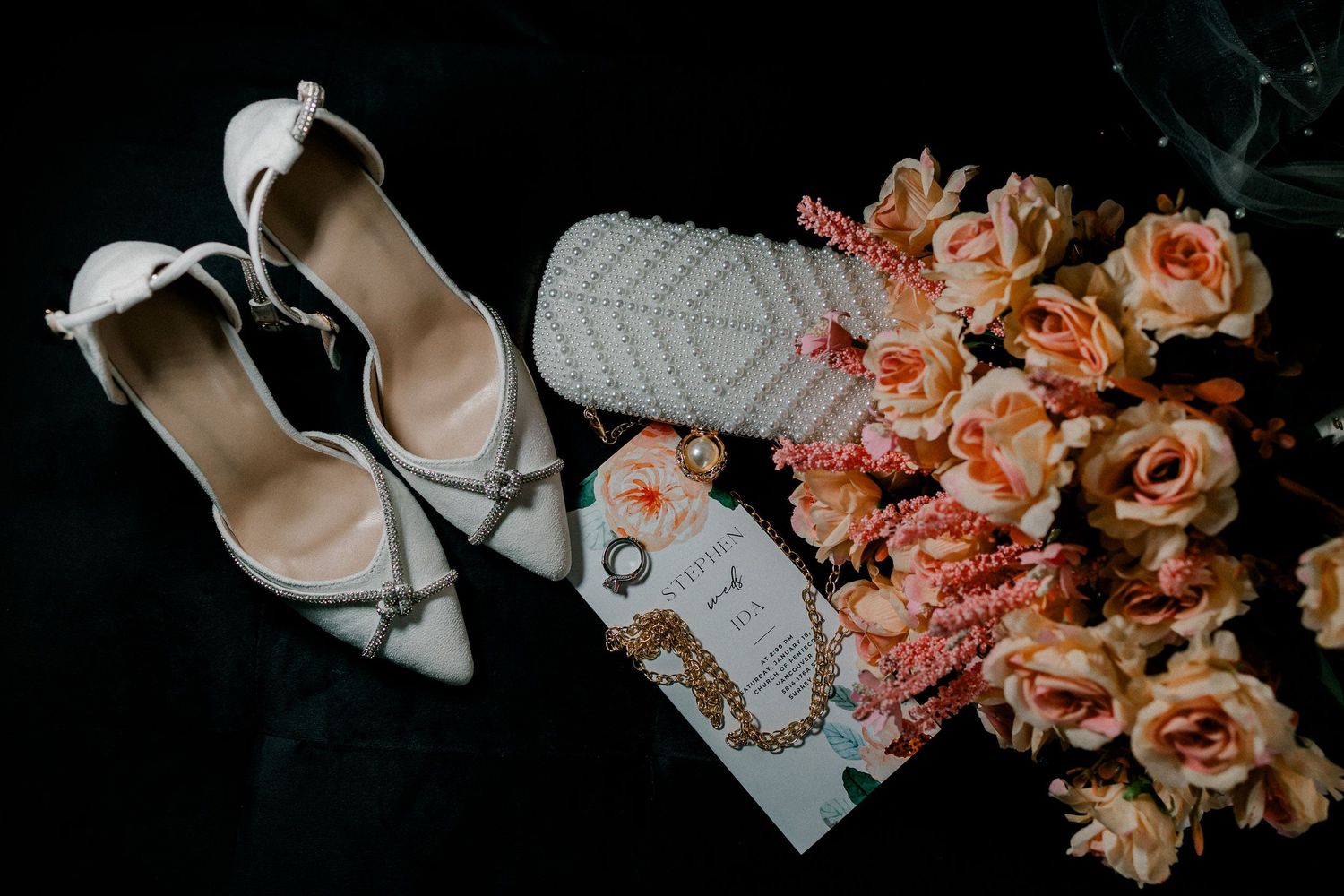 Elegant wedding details featuring white shoes, pink and peach roses, and delicate accessories arranged on a light surface.