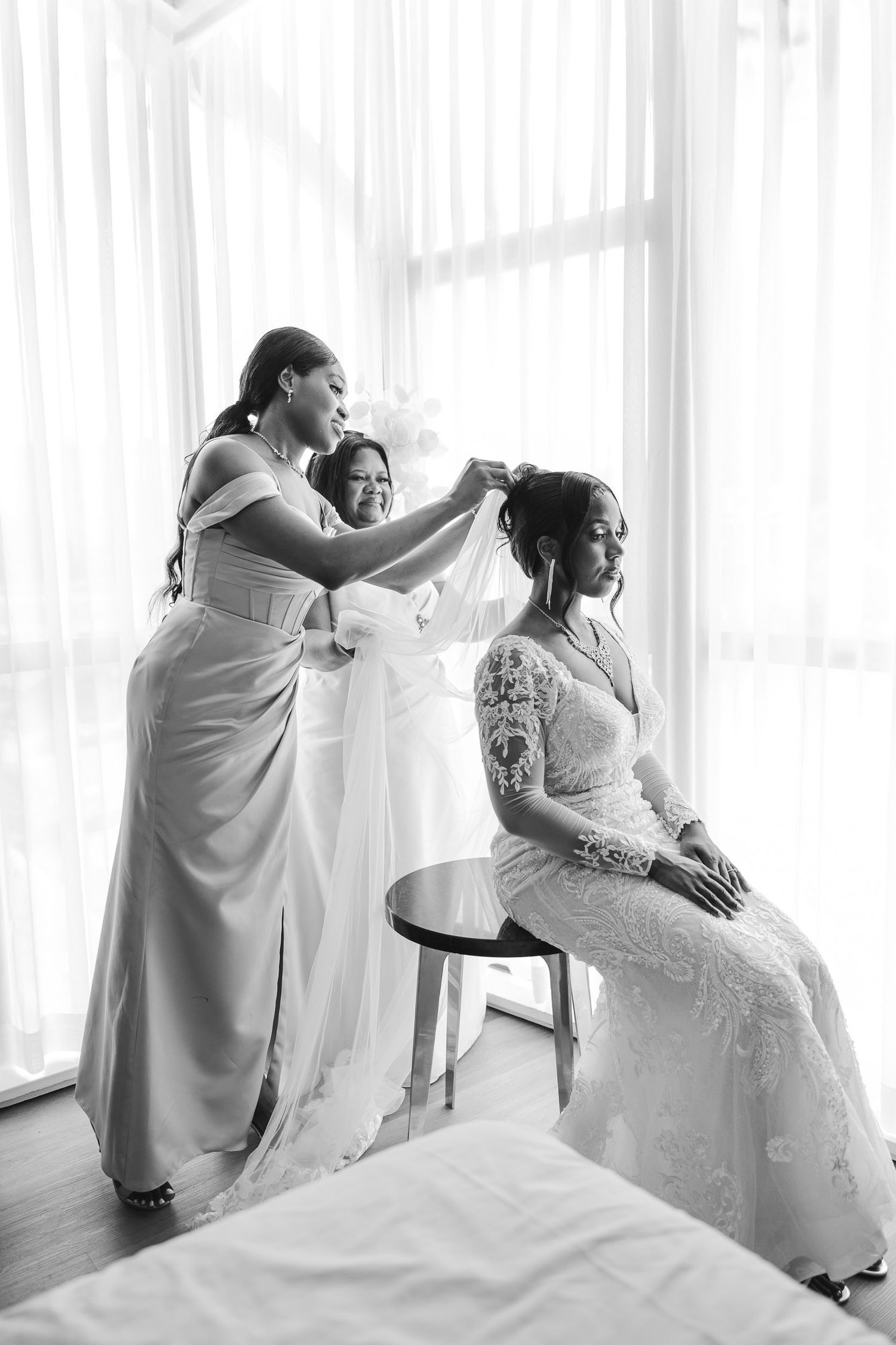 A series of black and white photos showing a bride in a lace wedding dress being helped with final preparations.