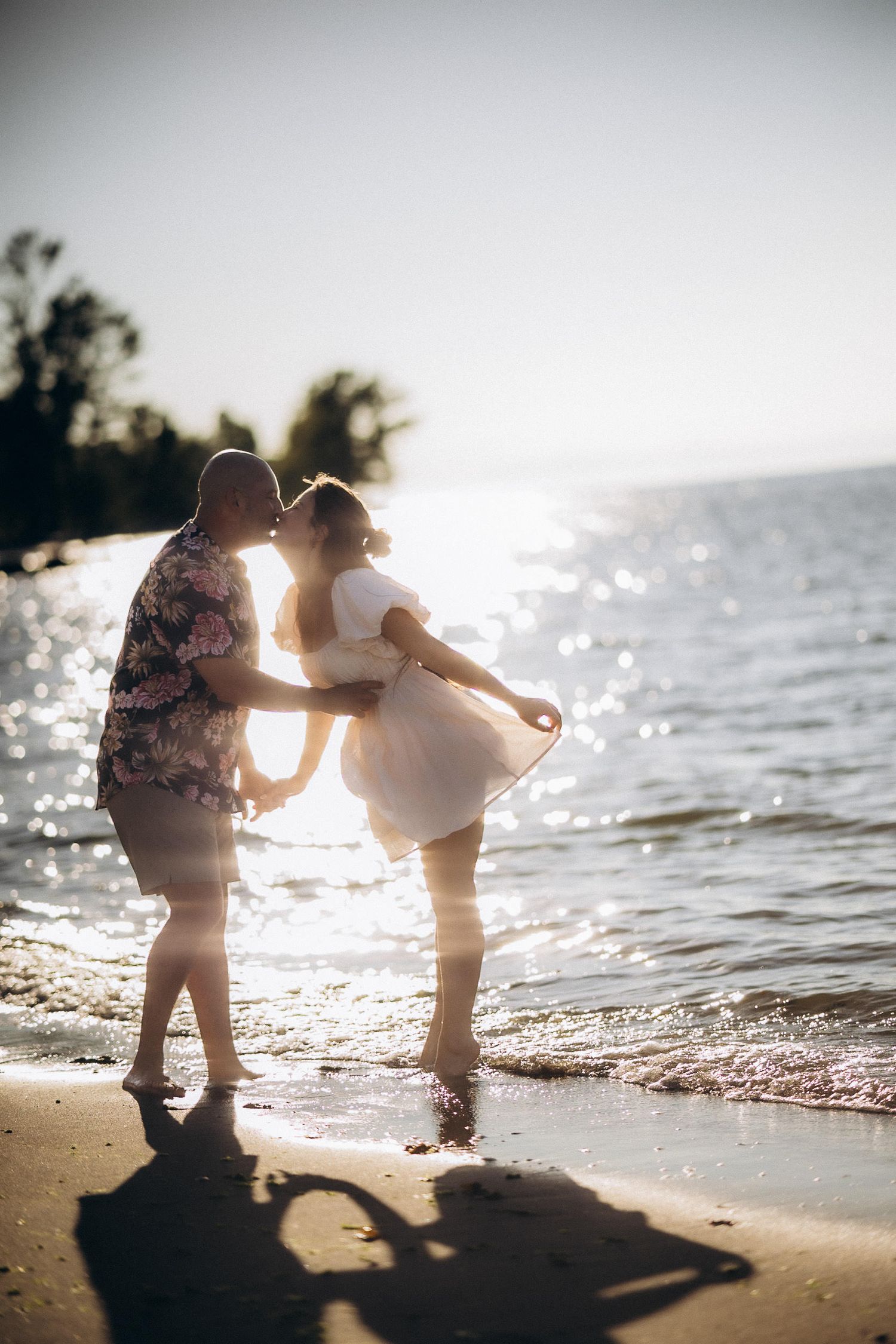 Two silhouettes dance together on a sunlit beach at golden hour as waves gently lap at the shoreline.