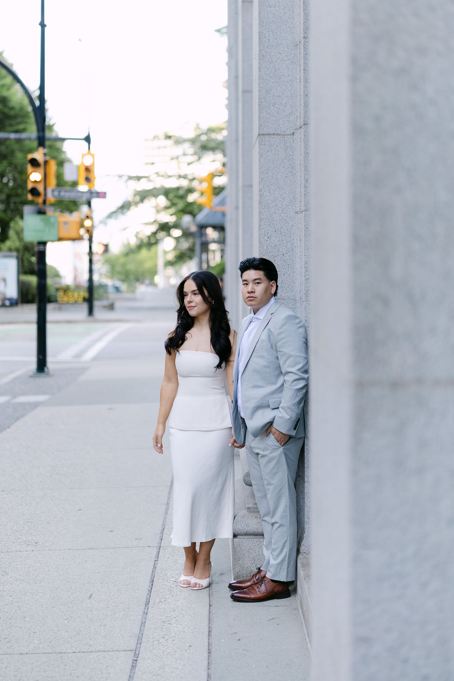 Couple poses against a granite building on a downtown sidewalk, she in a white dress and he in a light gray suit.