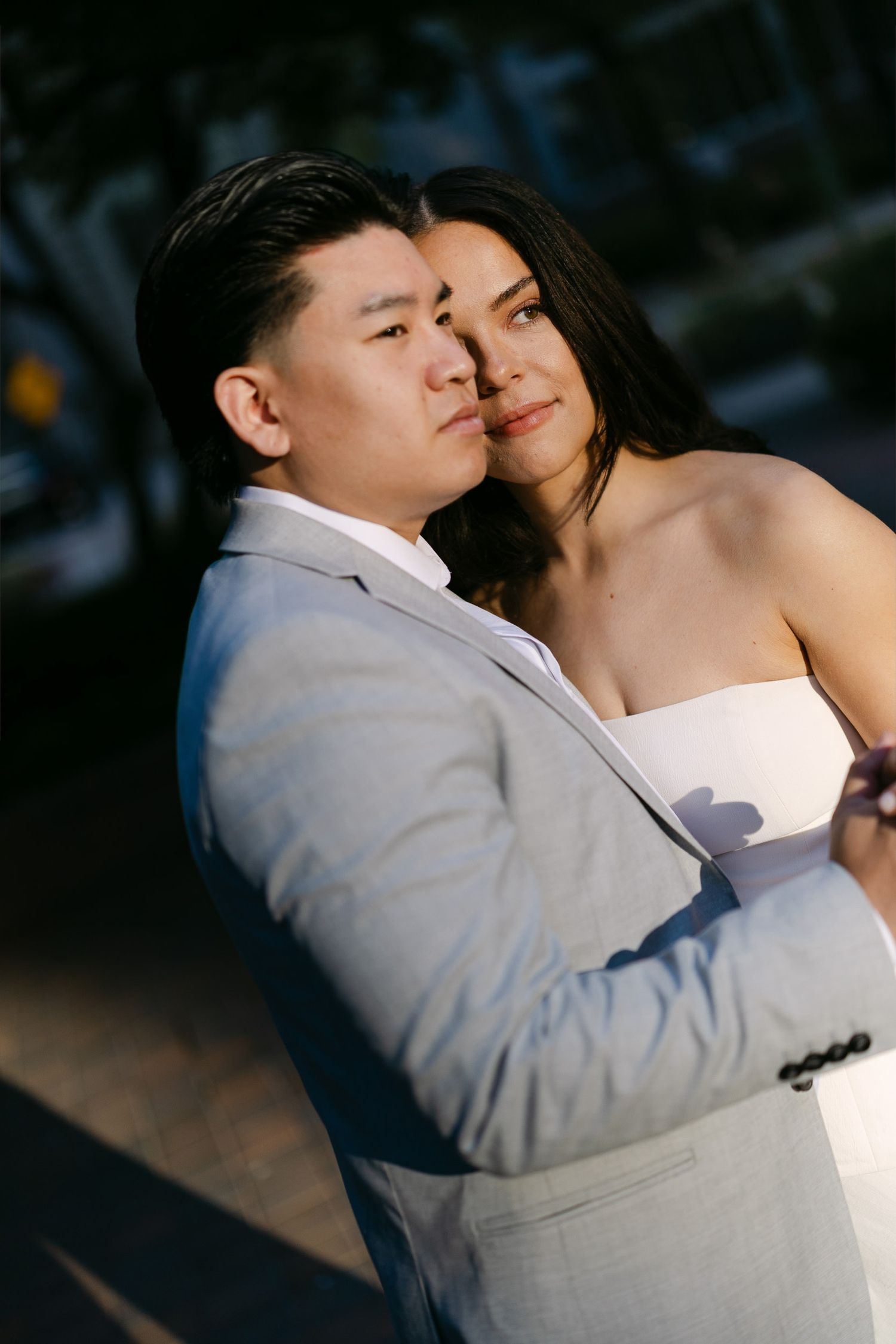Couple pose close together in warm sunlight, the man in a gray suit and the woman in a white strapless dress.