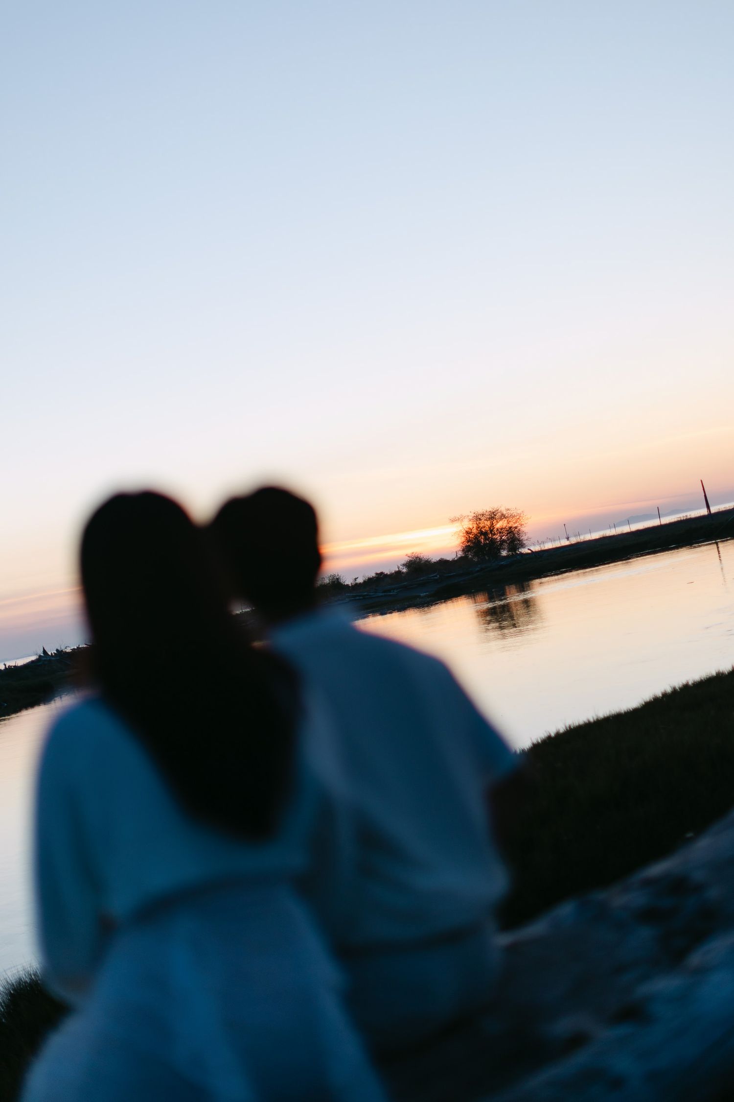 Blurred silhouette of a couple sitting close together by calm water at sunset, with a soft orange and pink sky.