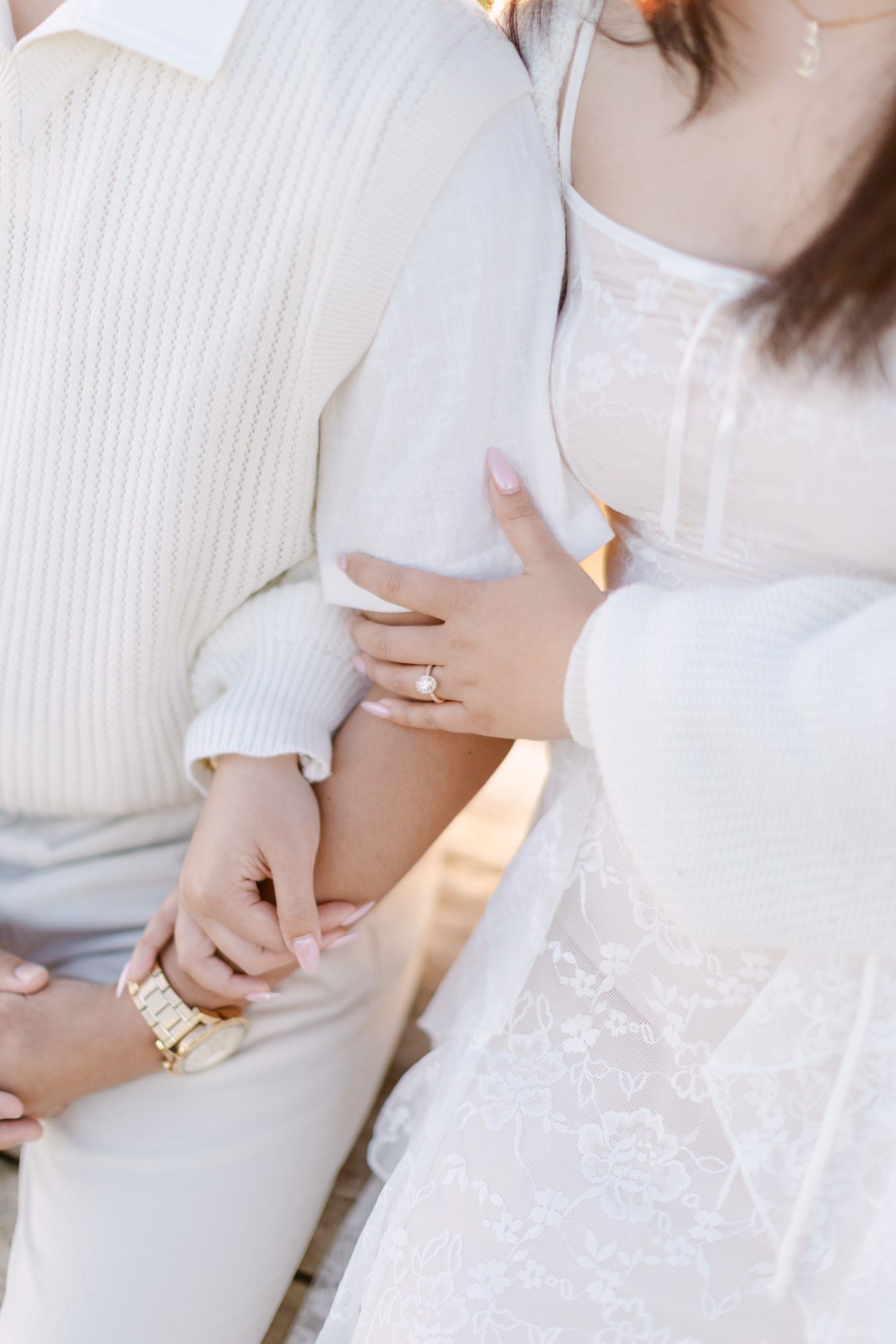 Close-up of a woman in a white lace dress with a diamond engagement ring resting her hand on her partner's arm.