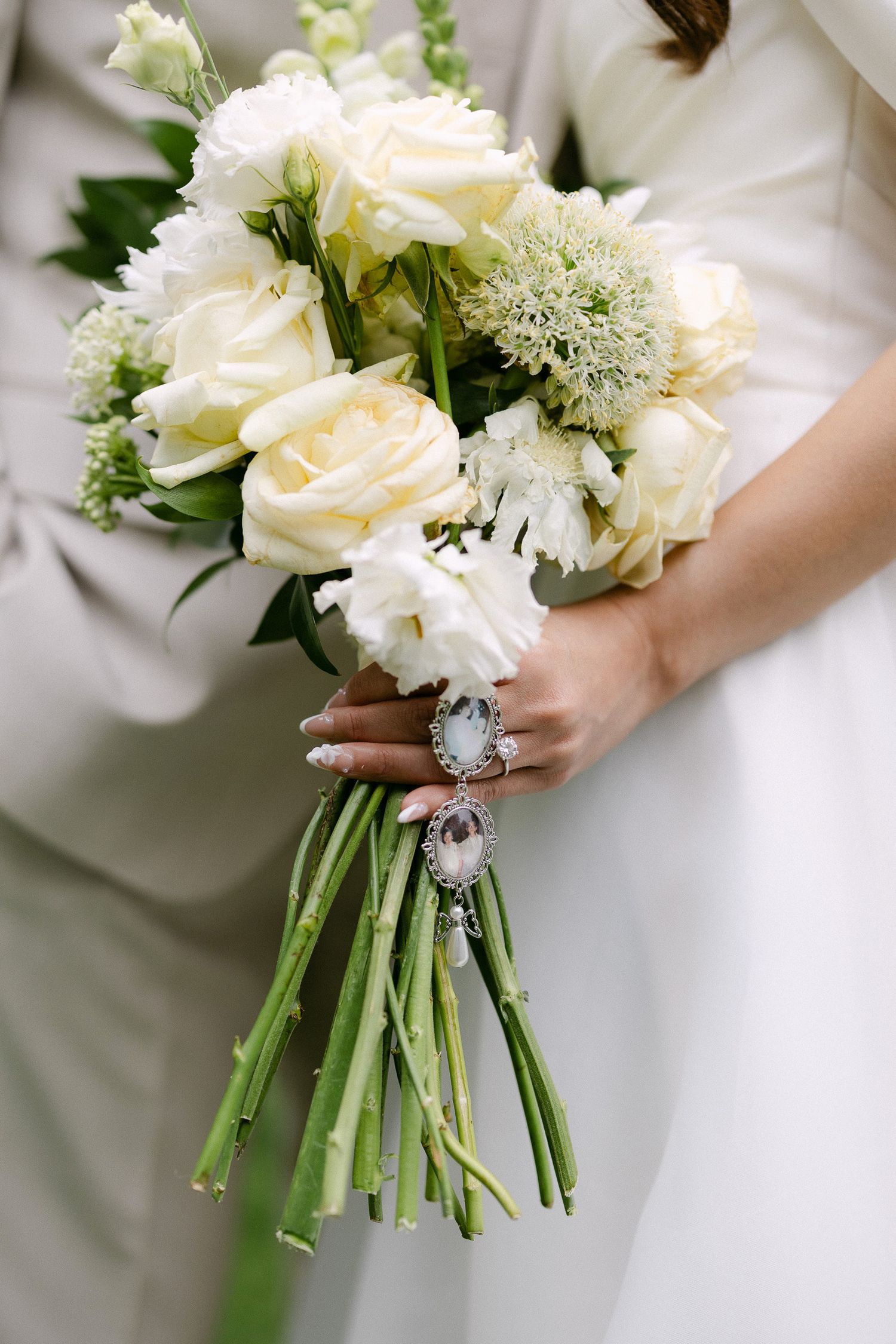 Bride holding white rose and green bouquet with silver brooch charm, wearing white gown beside groom in gray suit.