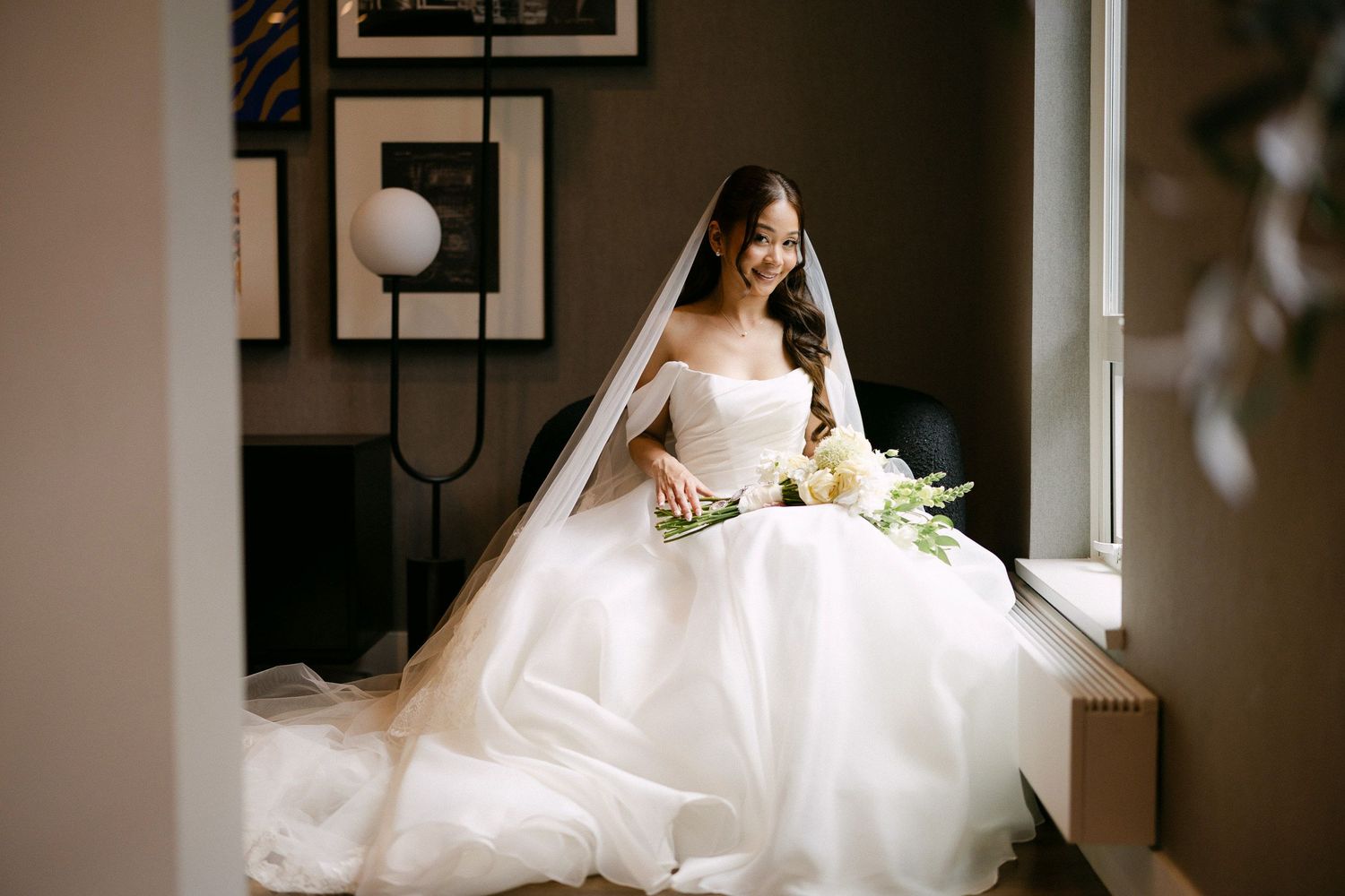 Bride in a white ball gown wedding dress with veil sits by a window holding a bouquet of white flowers.