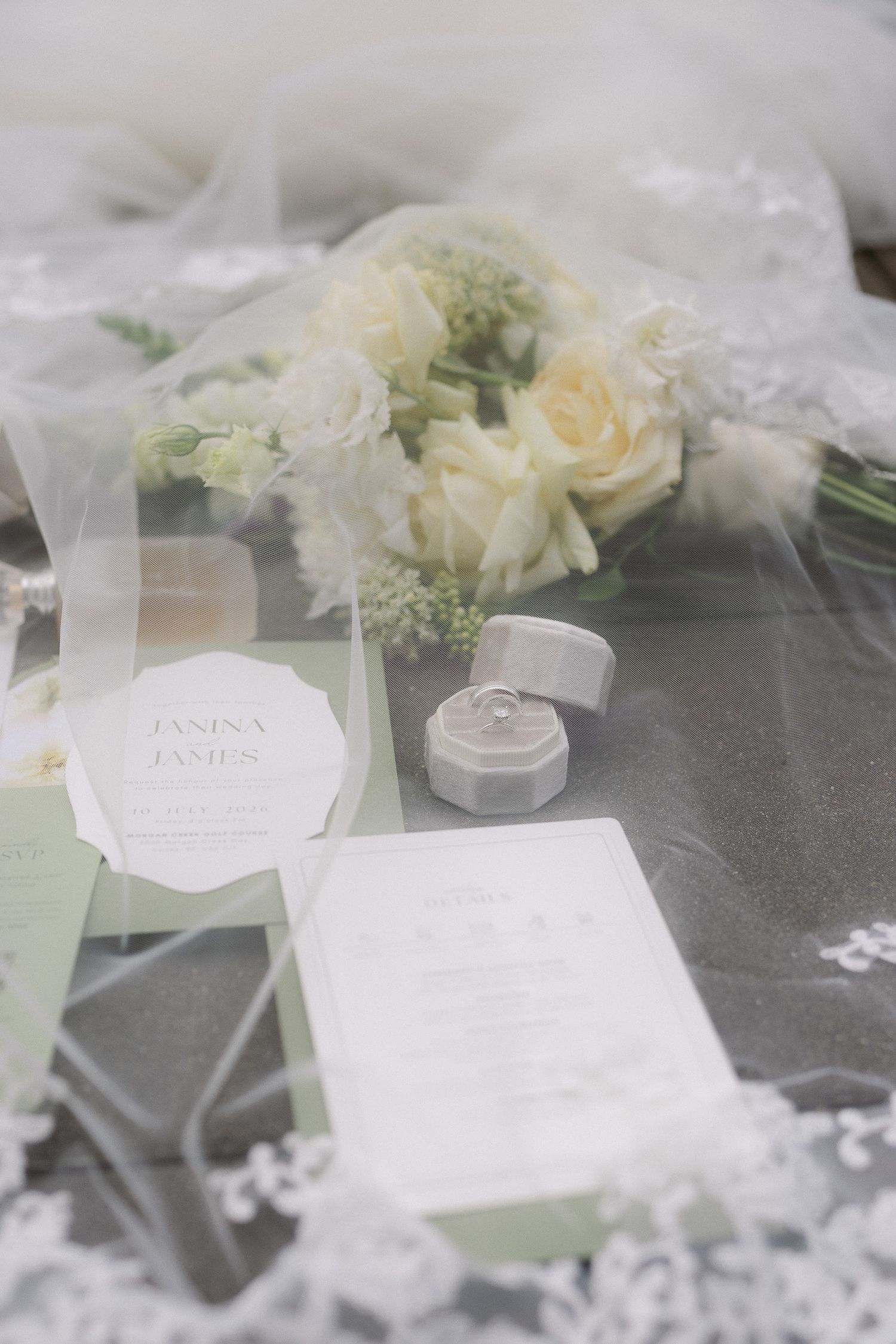Elegant wedding flat lay with white rose bouquet, ring box, and invitation suite draped in sheer veil on gray surface.