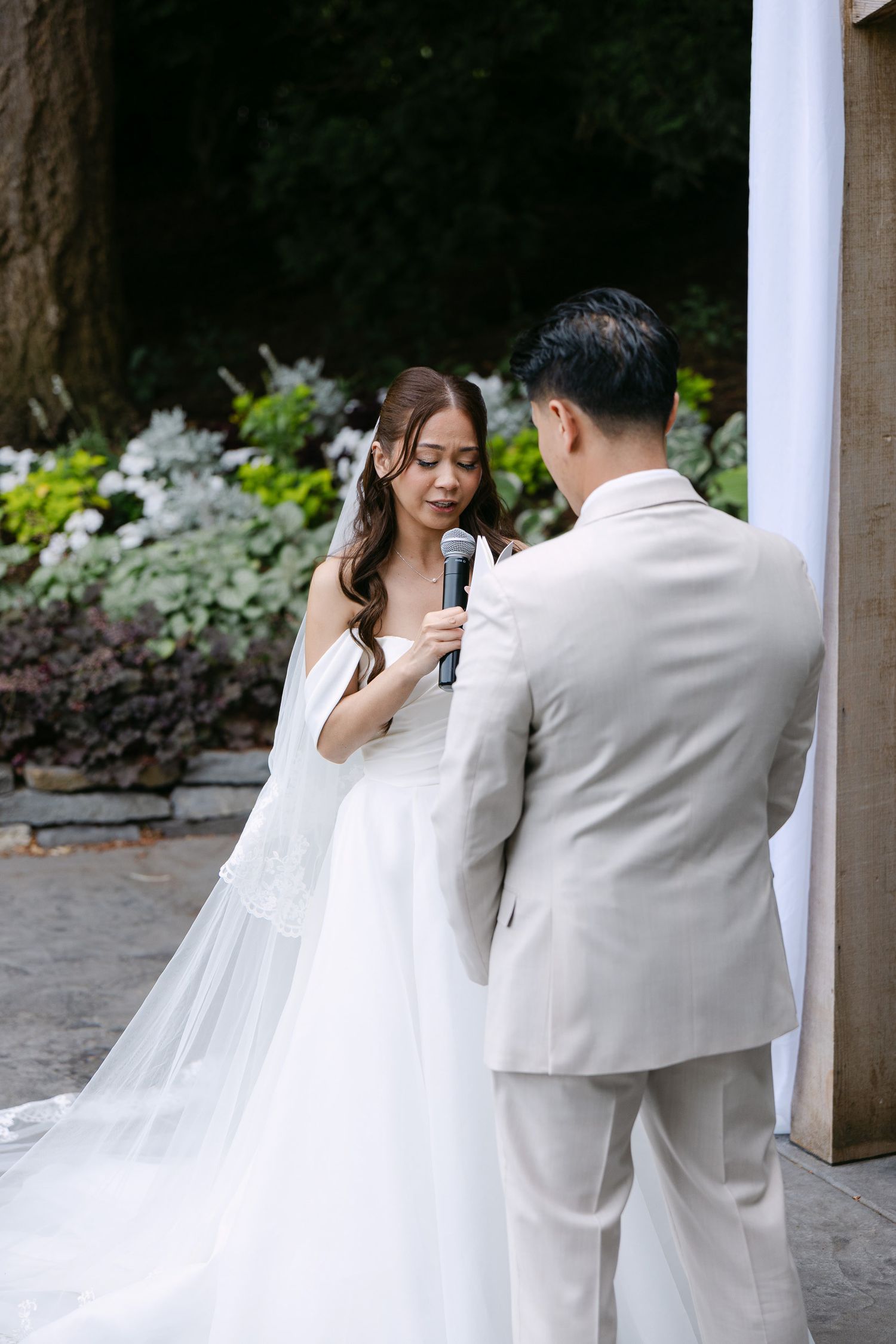 Bride reads wedding vows into microphone while groom listens during outdoor garden ceremony.