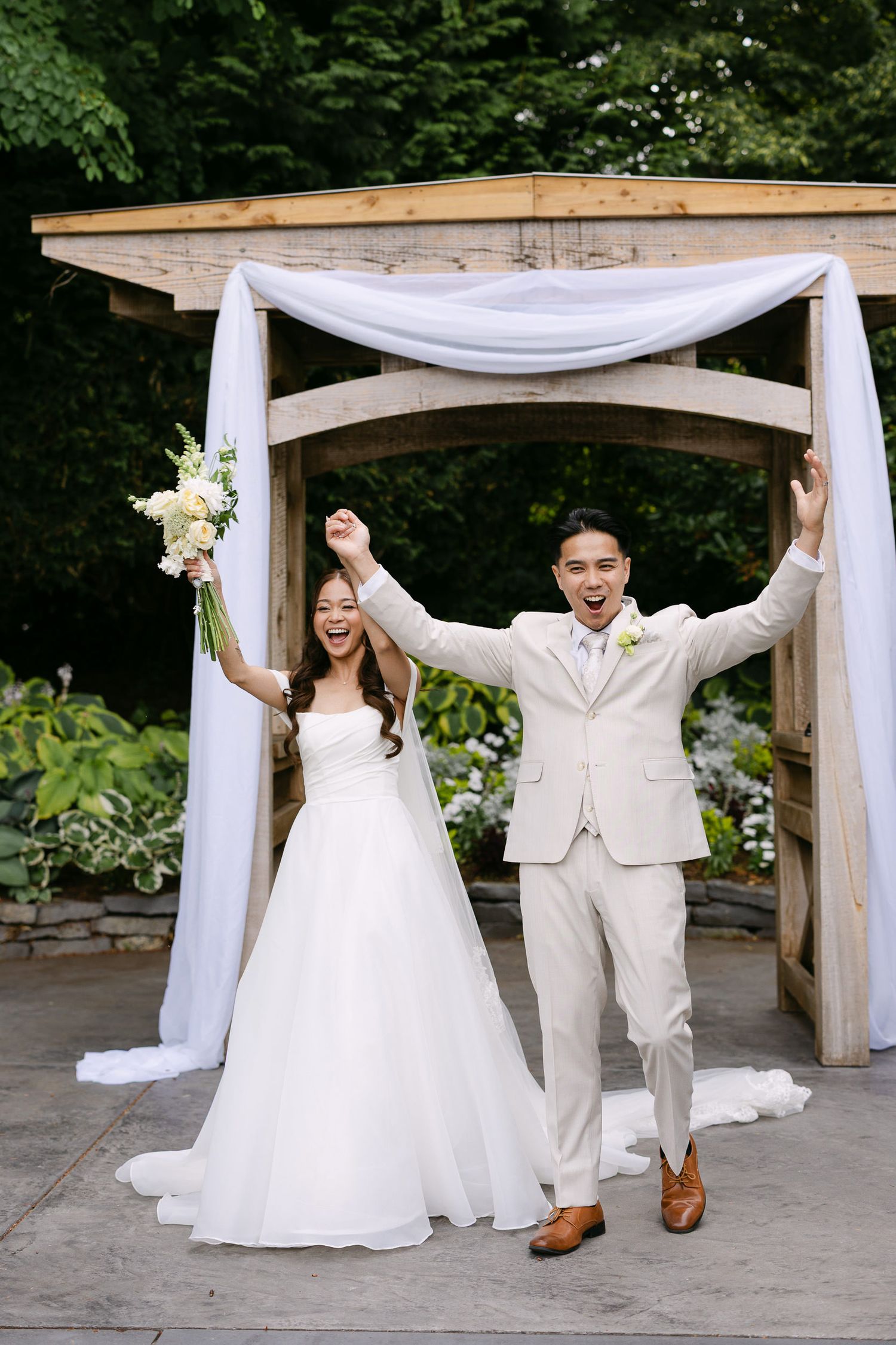Joyful newlywed couple celebrating under a white-draped wooden arch, bride in white gown holding bouquet, groom in beige suit.