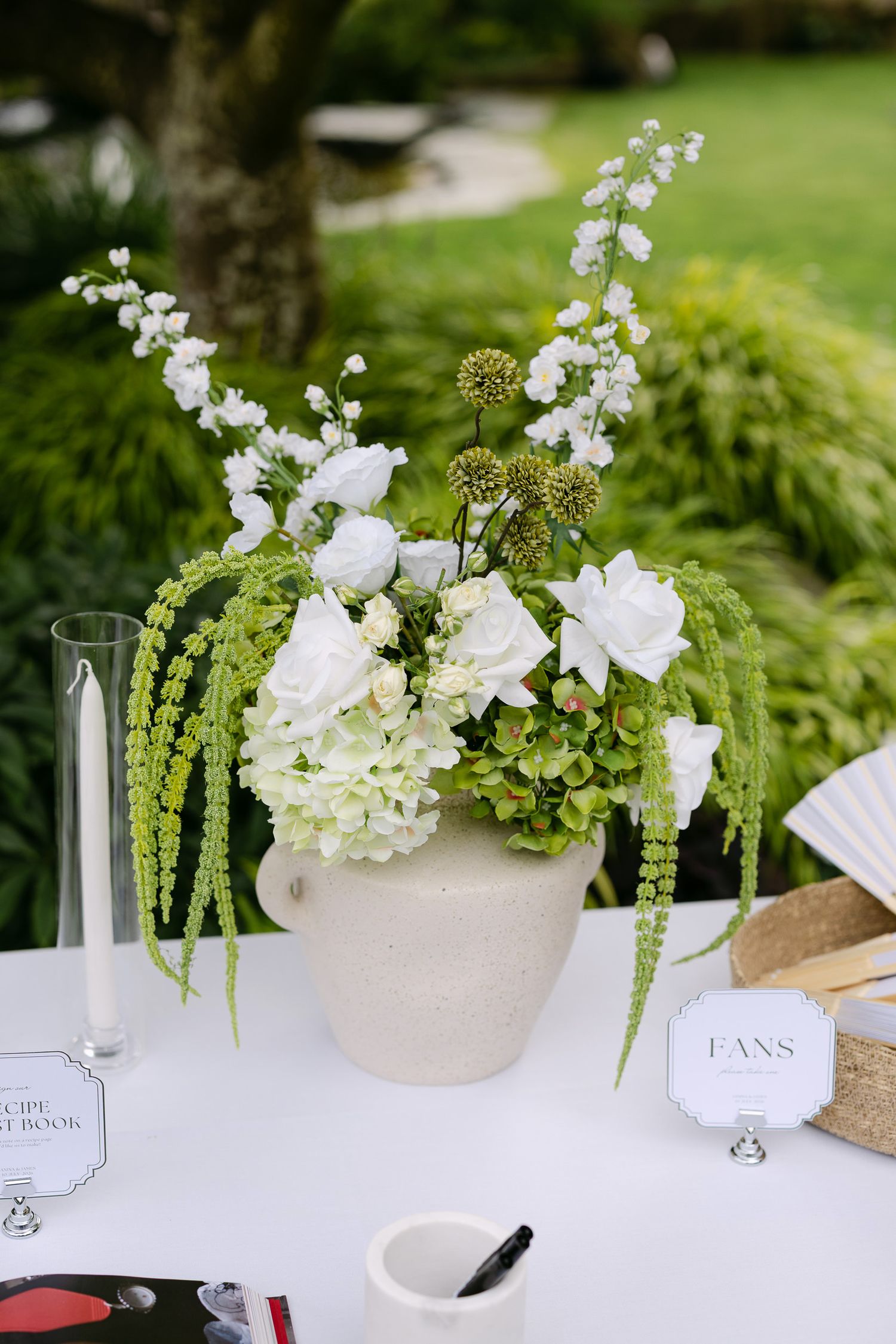 White floral centerpiece in ceramic mug with green accents on white table at outdoor wedding reception.