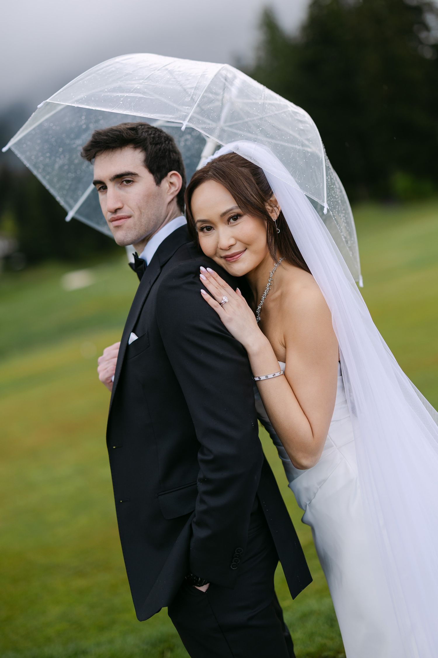 Bride and groom pose under a clear bubble umbrella on a lush green lawn on their rainy wedding day.