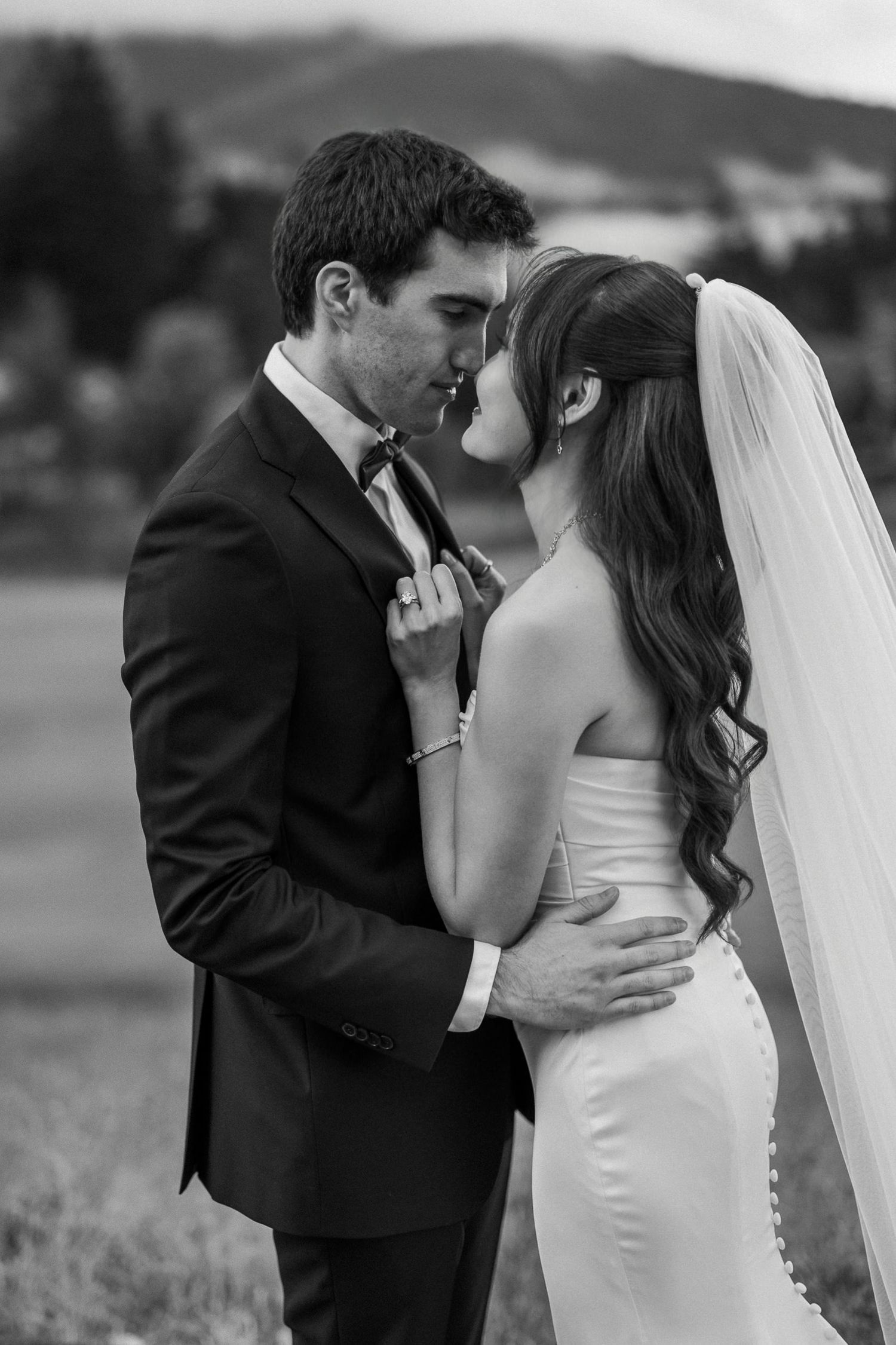Black and white photo of a bride and groom sharing a romantic kiss outdoors with rolling hills in the background.