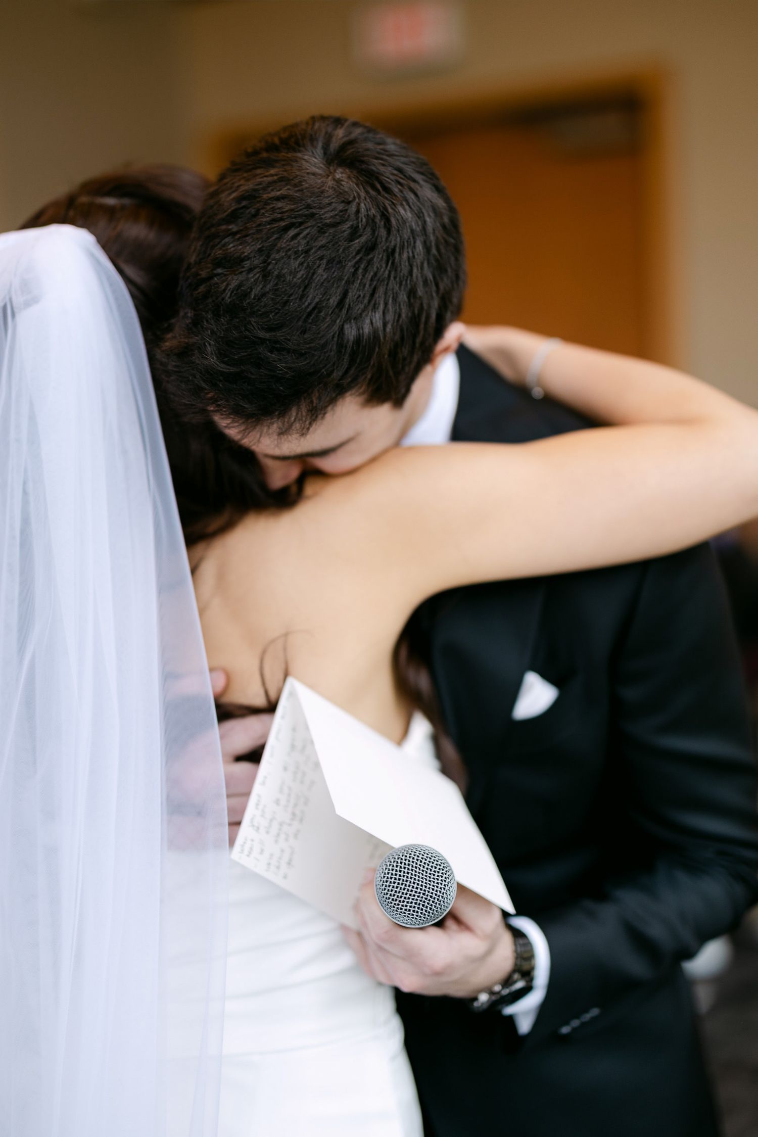 Bride and groom sharing an emotional embrace during wedding ceremony, groom holding microphone and paper vows.