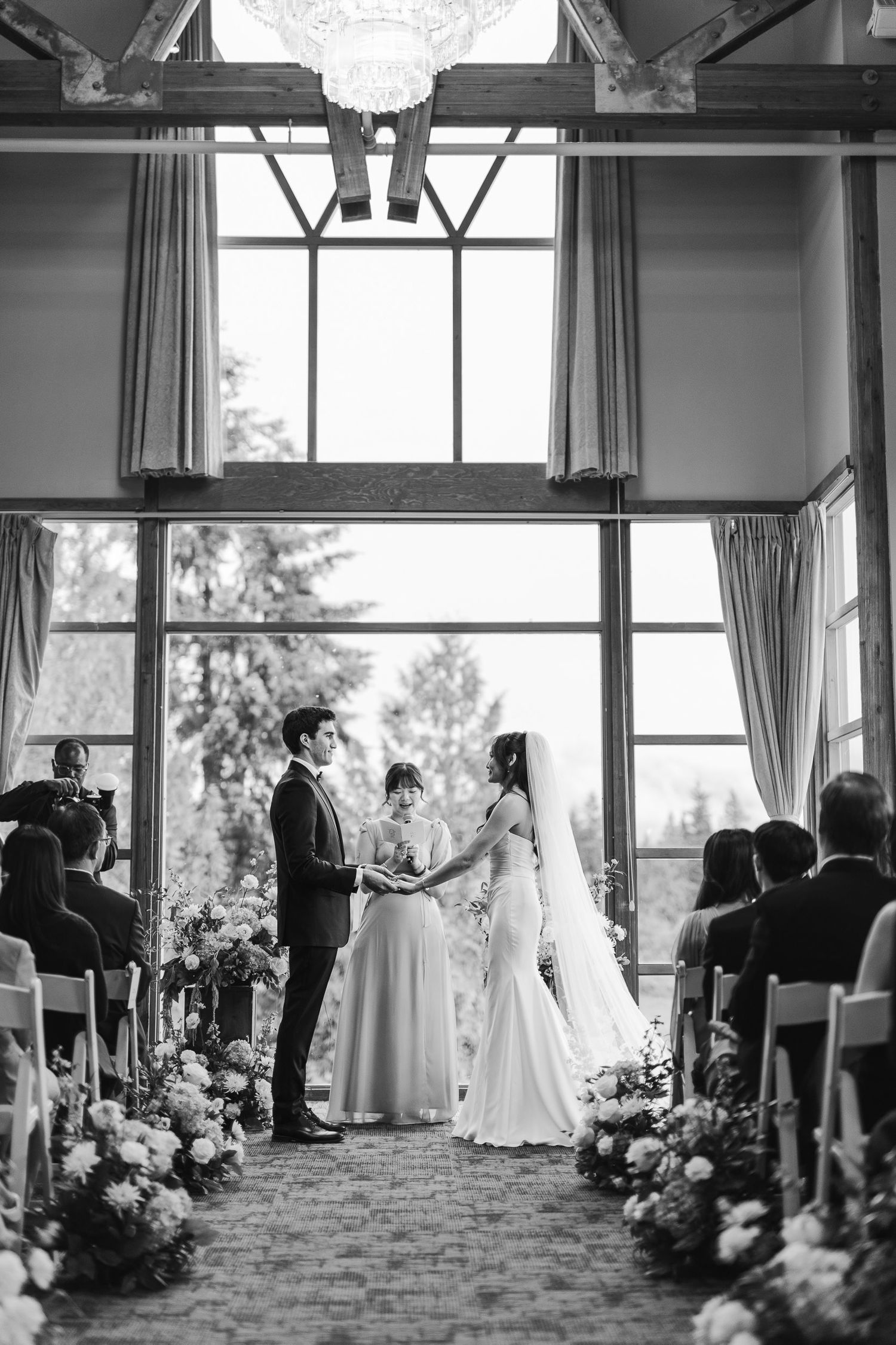 Black and white wedding ceremony photo of bride and groom holding hands at the altar with floral arrangements and large windows.
