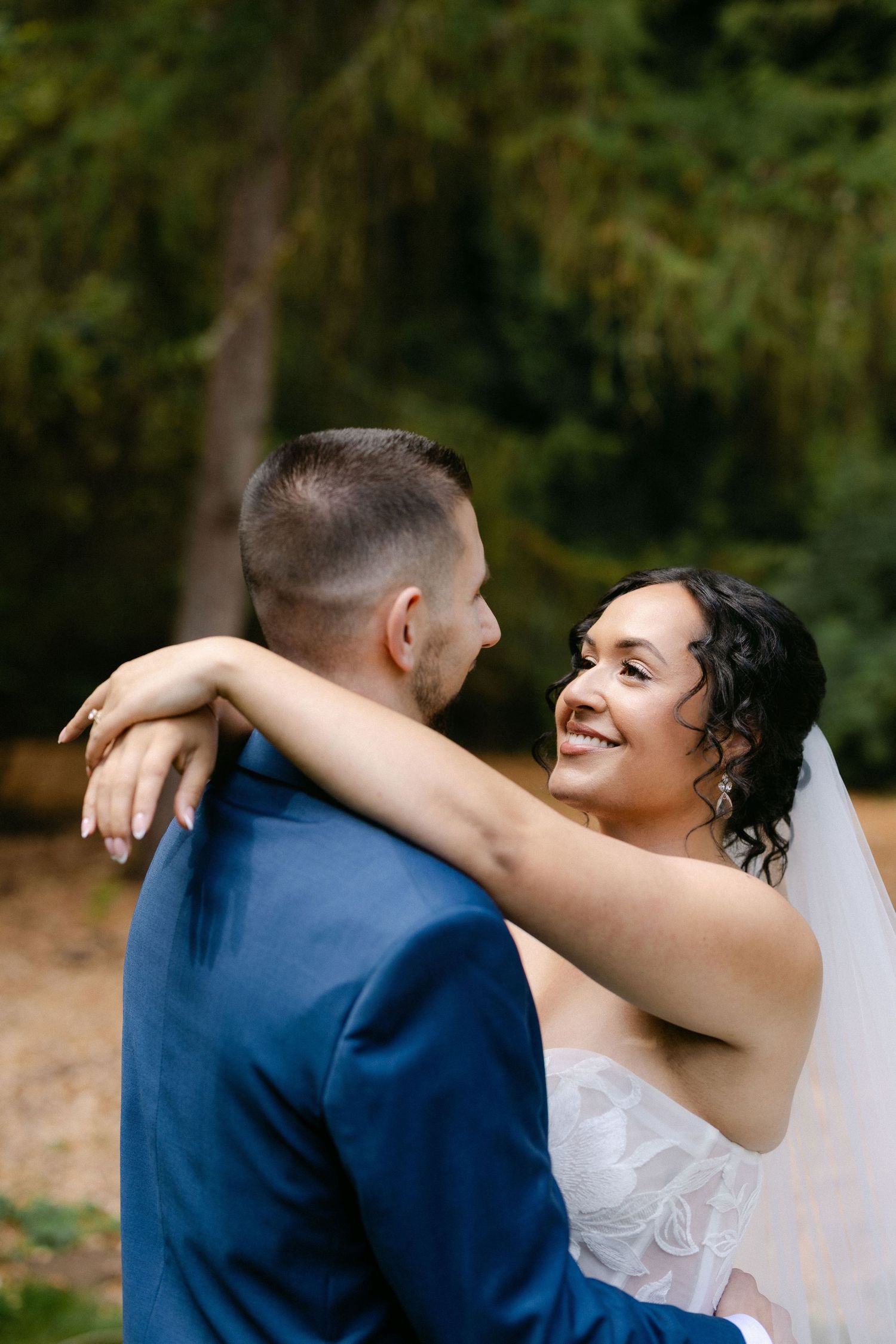 Smiling bride in white lace dress embraces groom in blue suit outdoors with green trees in background.
