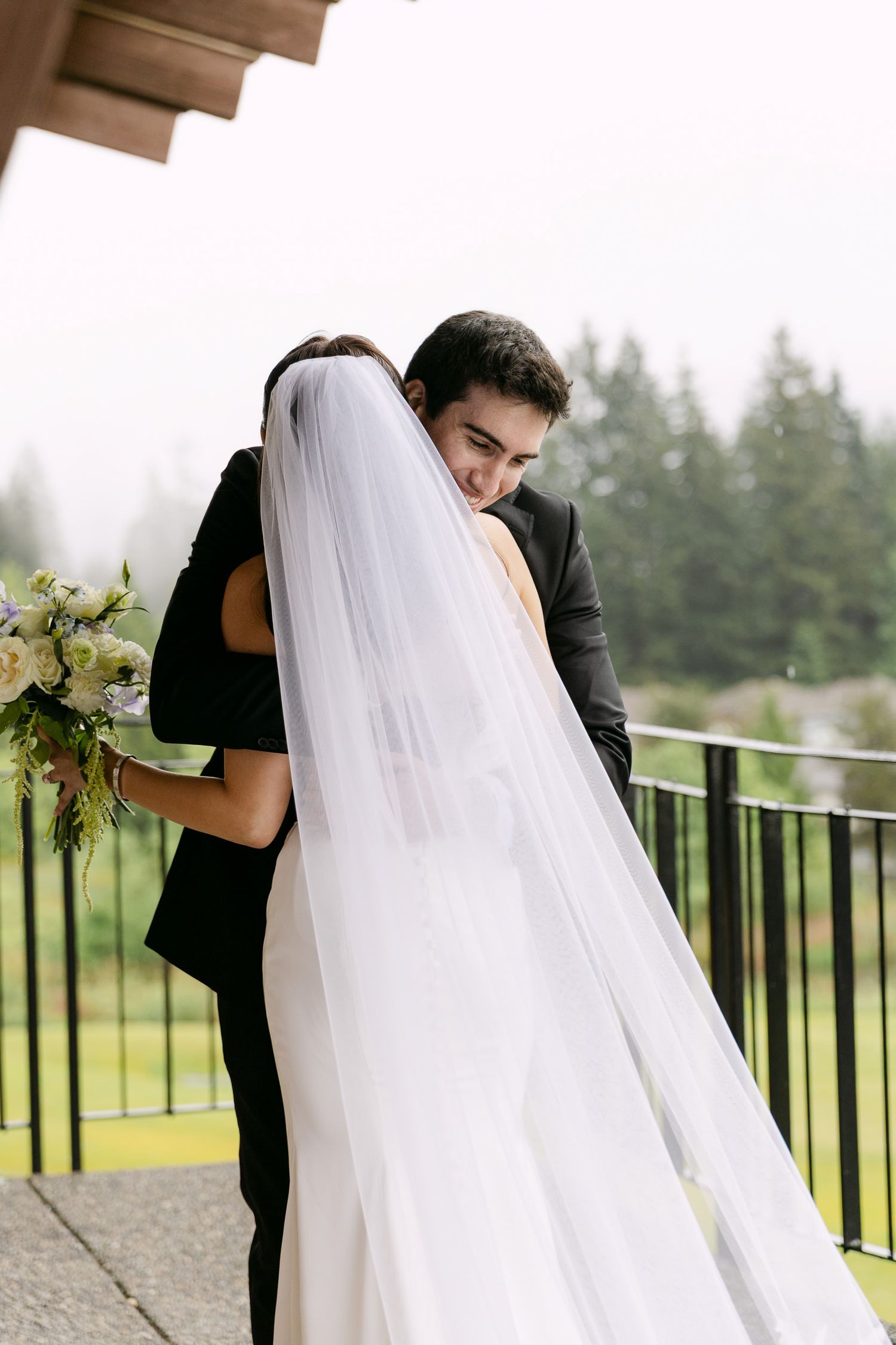 A groom in a black suit embraces his bride in a white gown and veil on a balcony, surrounded by lush greenery.