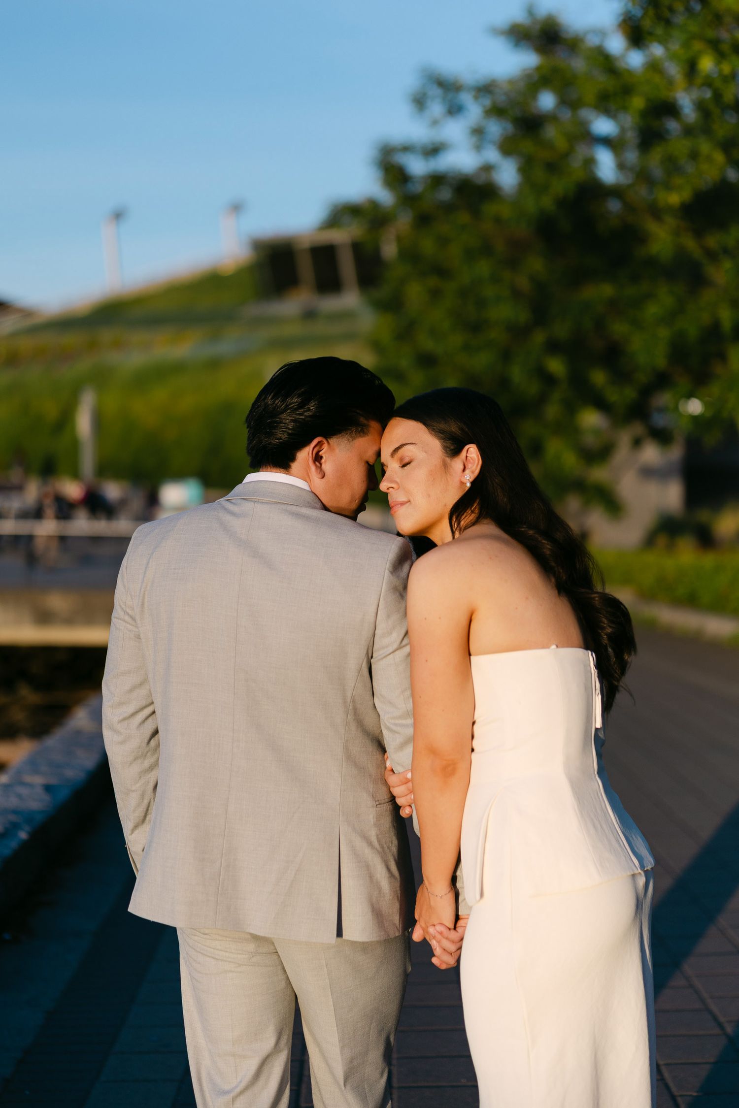 Couple in a gray suit and white strapless dress embracing with foreheads touching on a sunlit waterfront promenade at golden hour.