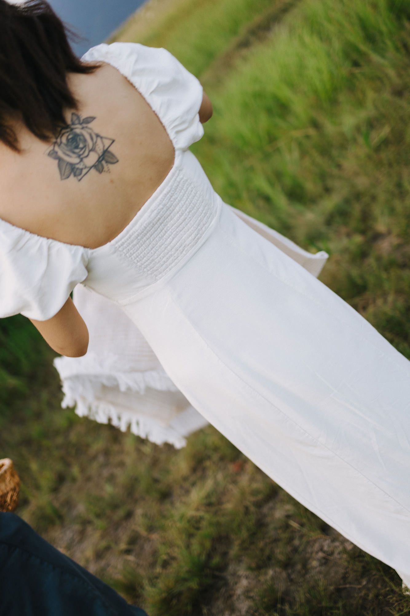Woman in a white off-shoulder smocked dress with a floral back tattoo stands on a grassy hillside.