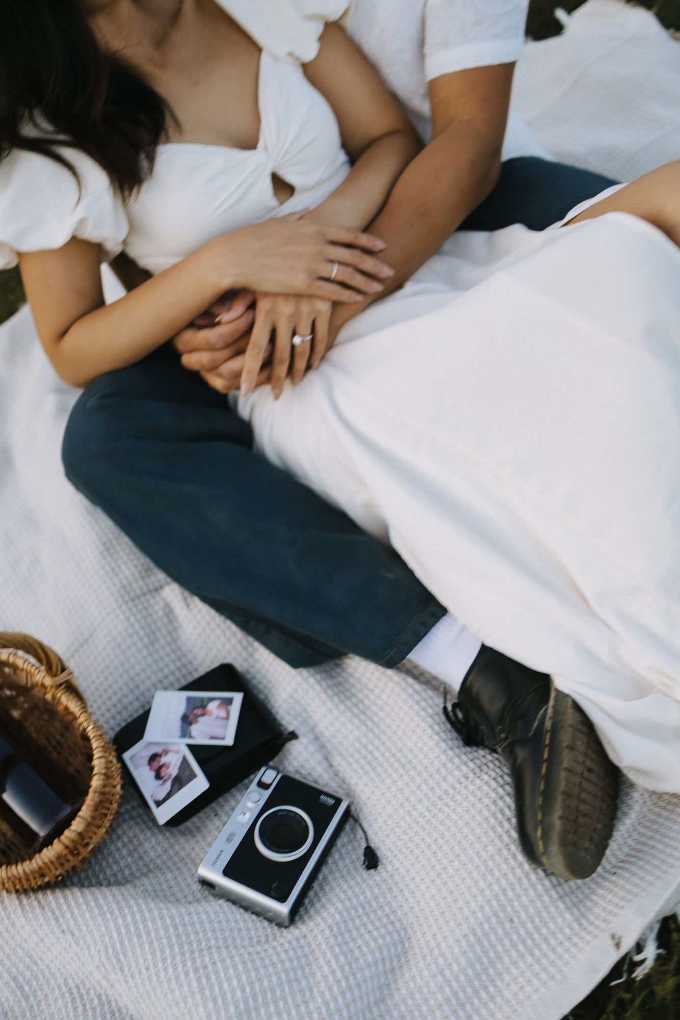 Couple in white and jeans sit close on a picnic blanket with hands clasped, a wedding ring, instant camera and wicker basket nearby.