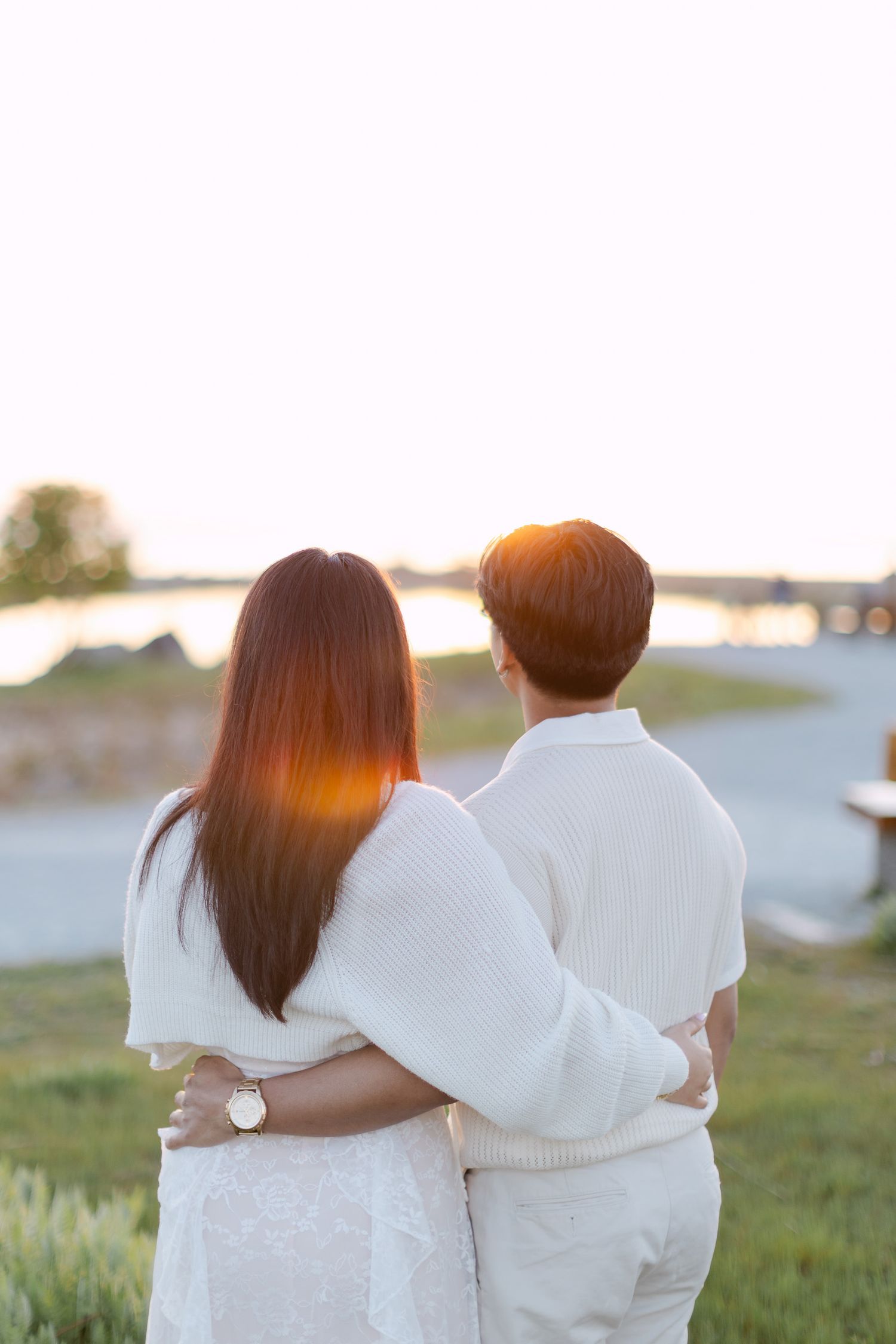 Couple in matching white outfits embracing from behind at sunset, looking out over a peaceful waterfront with warm golden light.