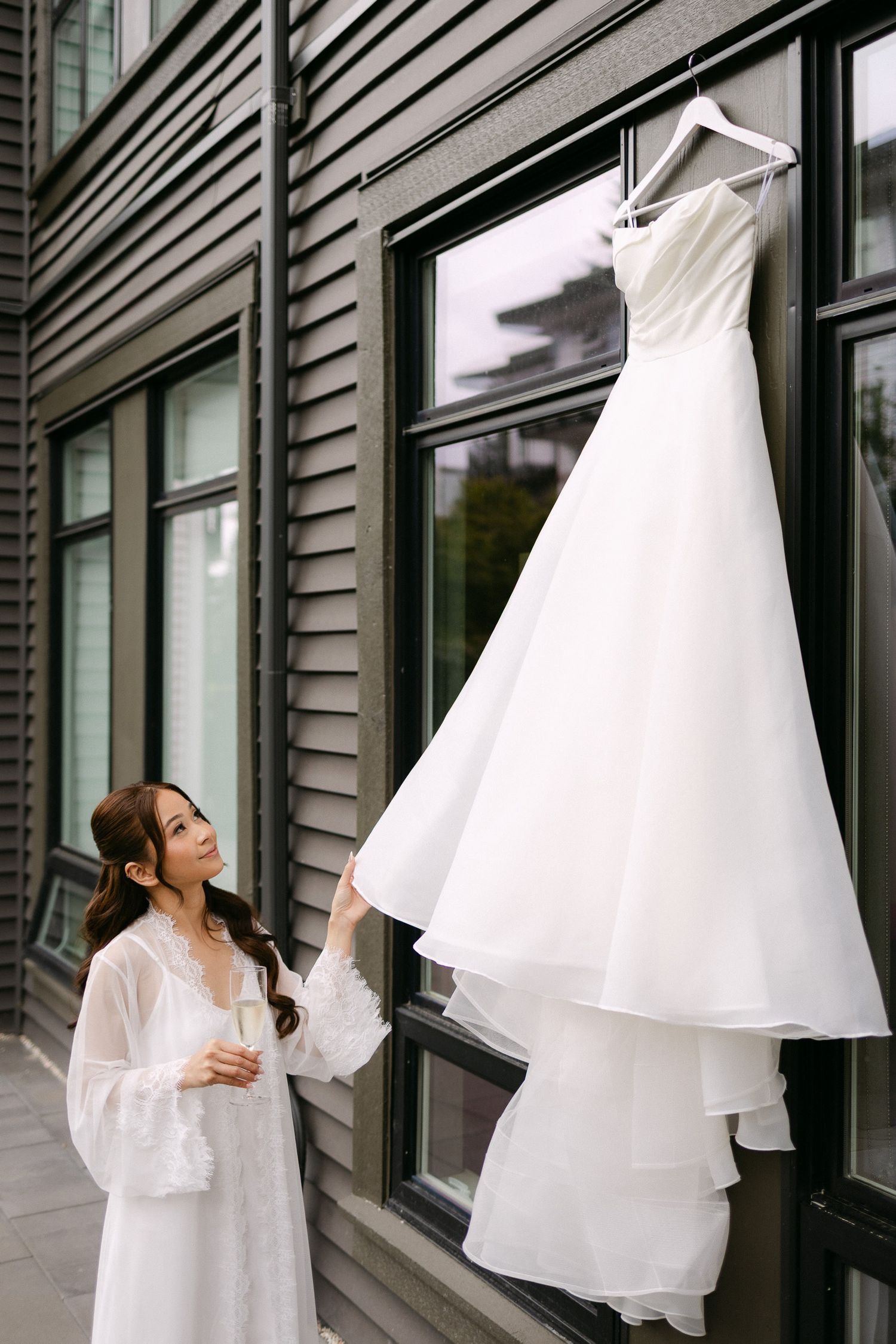 Bride in white robe admiring her elegant wedding gown hanging outside a modern building.